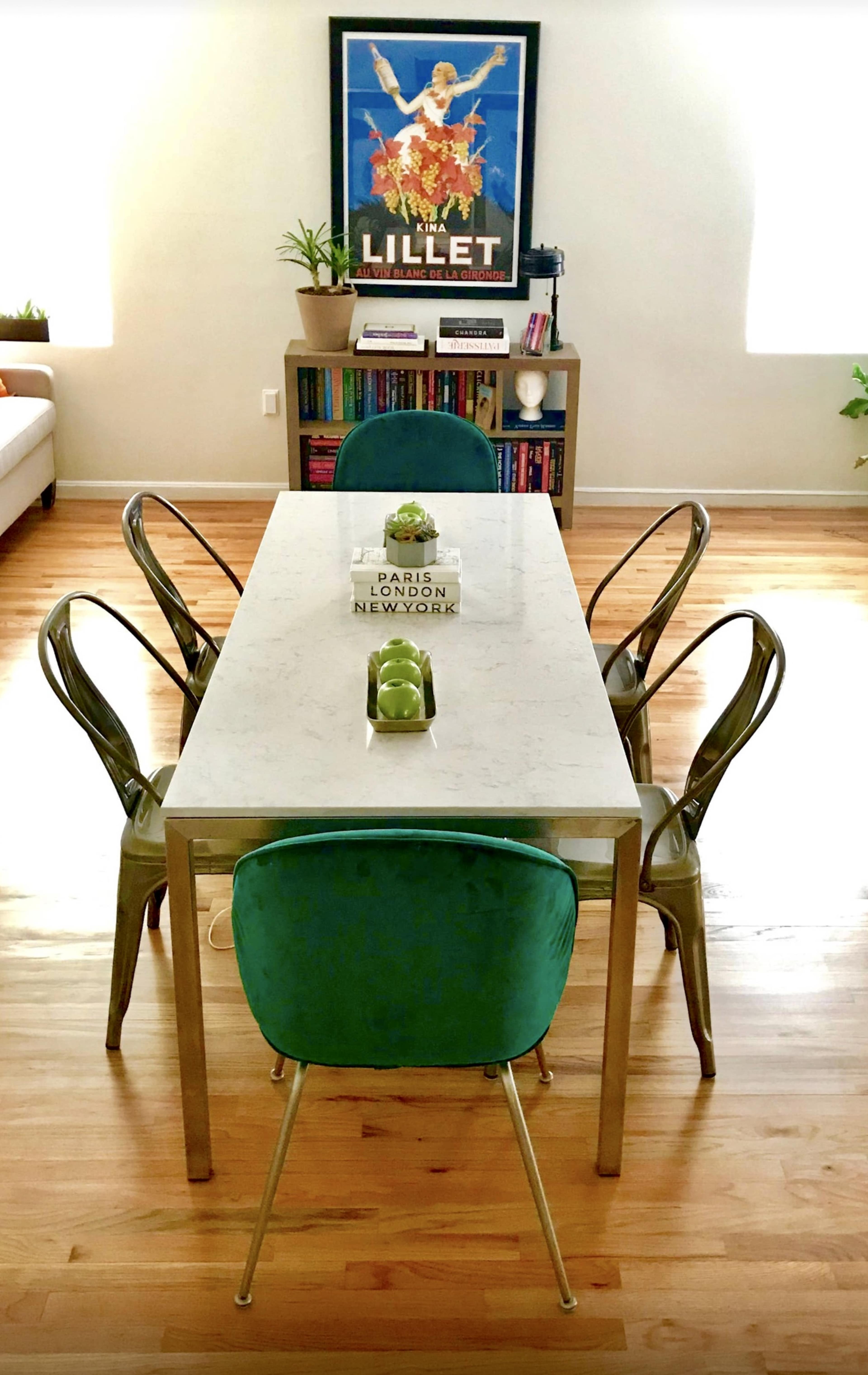 A modern dining table with four chairs is set in a bright room with a bookshelf and a framed poster in the background.