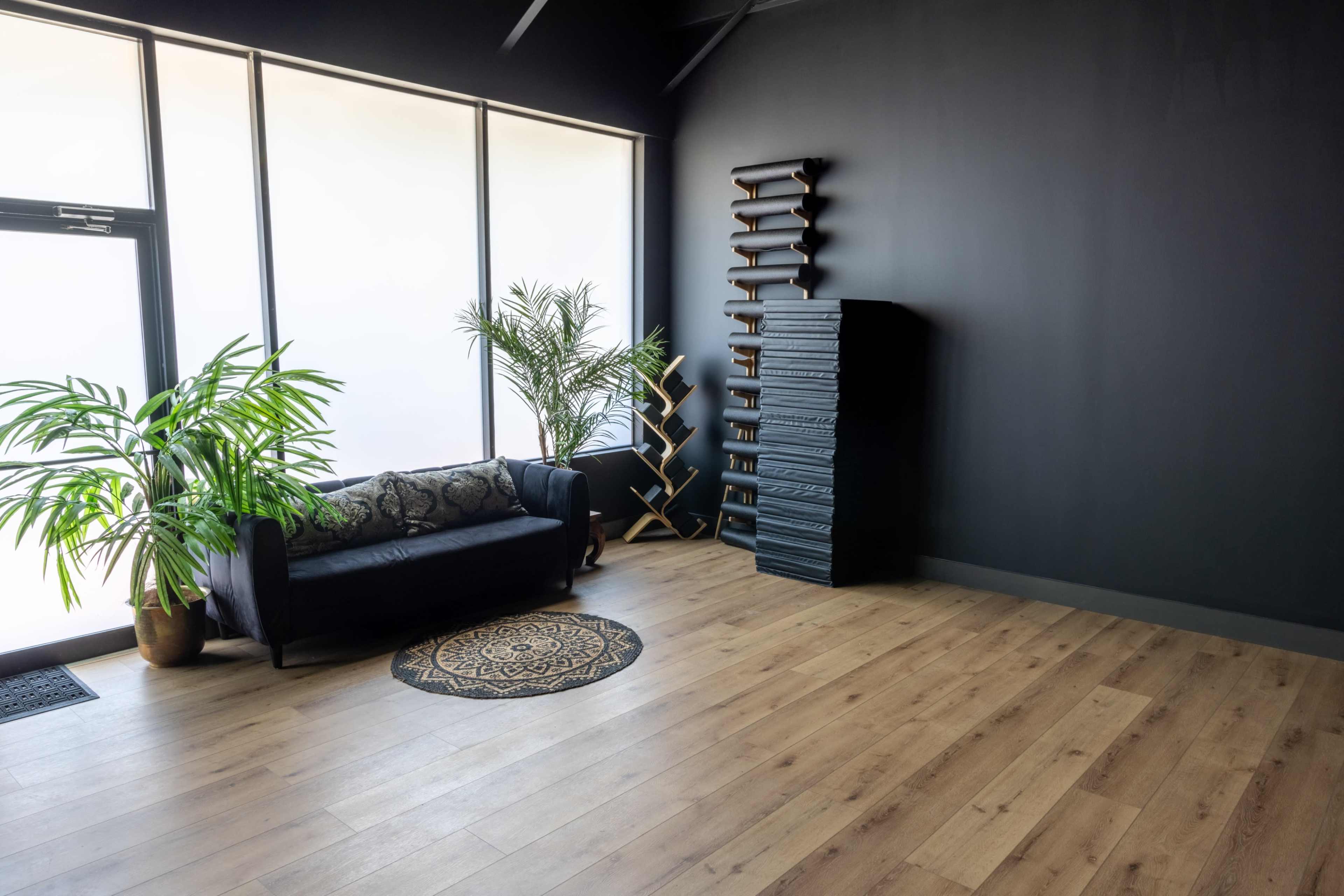 A spacious, minimally furnished room with a black wall, large windows, a dark couch, a decorative rug, and a tall wooden shelf.