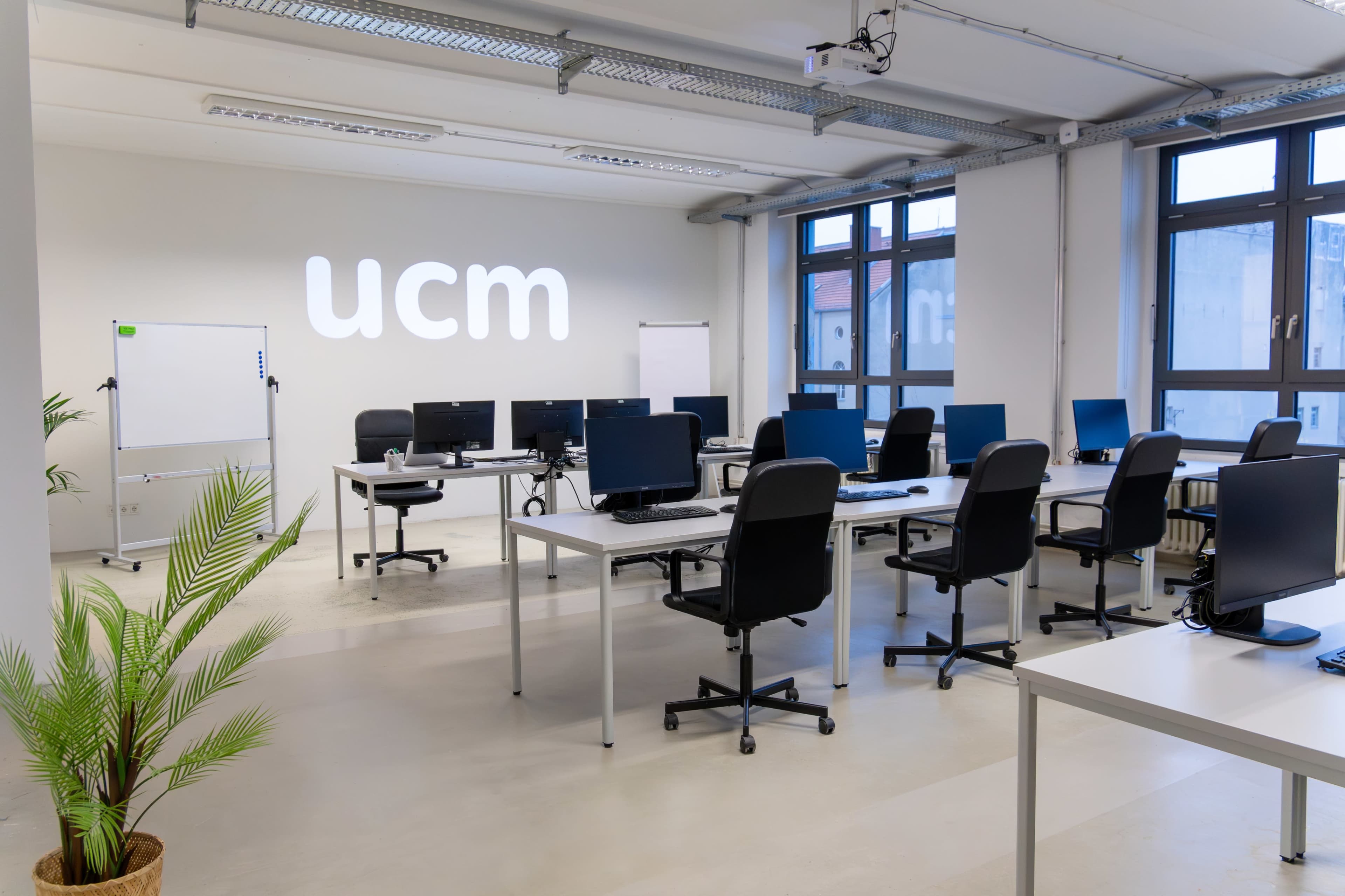 The image shows a modern office space with multiple black desks, computer monitors, and a large wall displaying the letters "ucm."