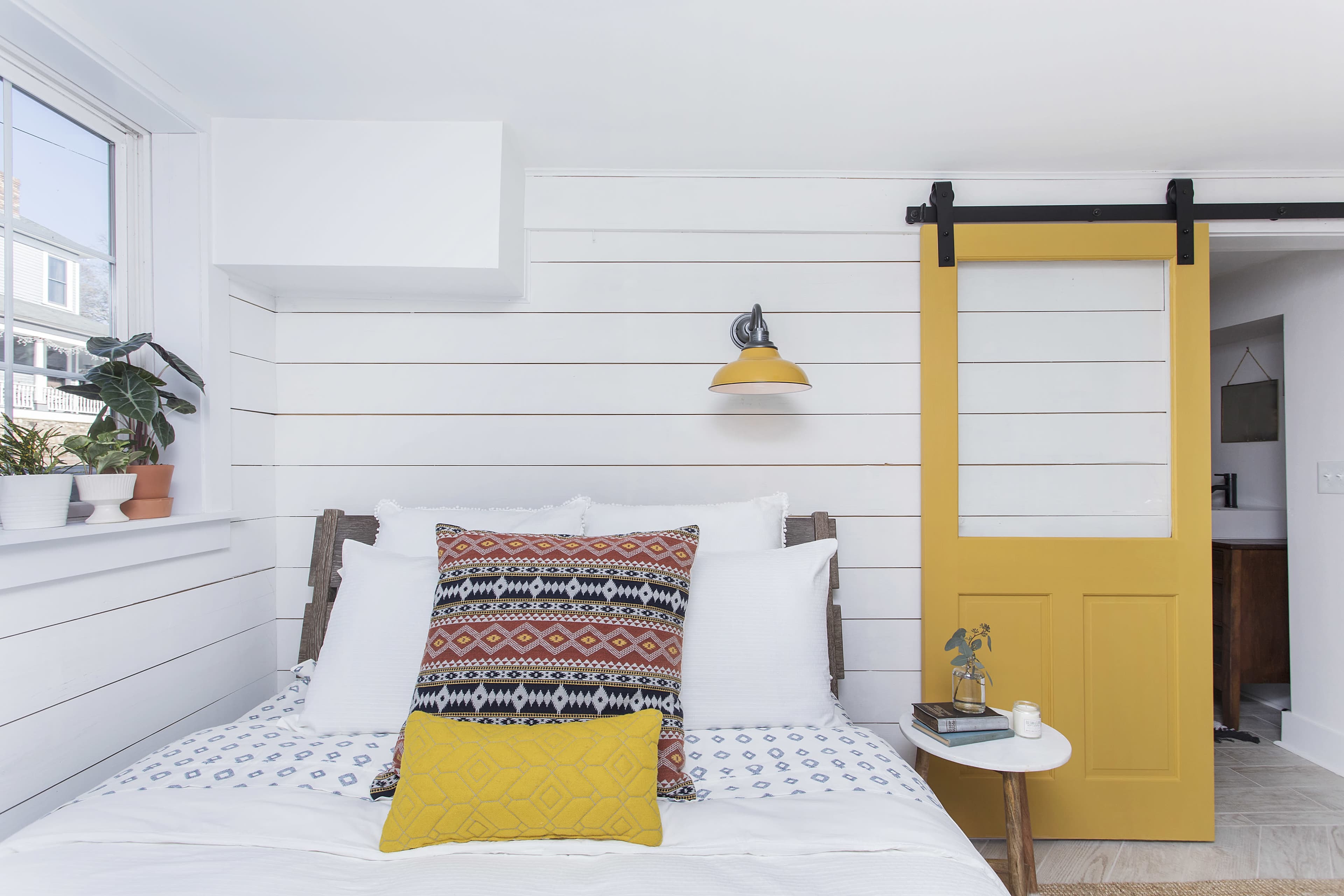 A bright, minimalist bedroom featuring a white bed with patterned pillows, a yellow sliding barn door, and indoor plants.