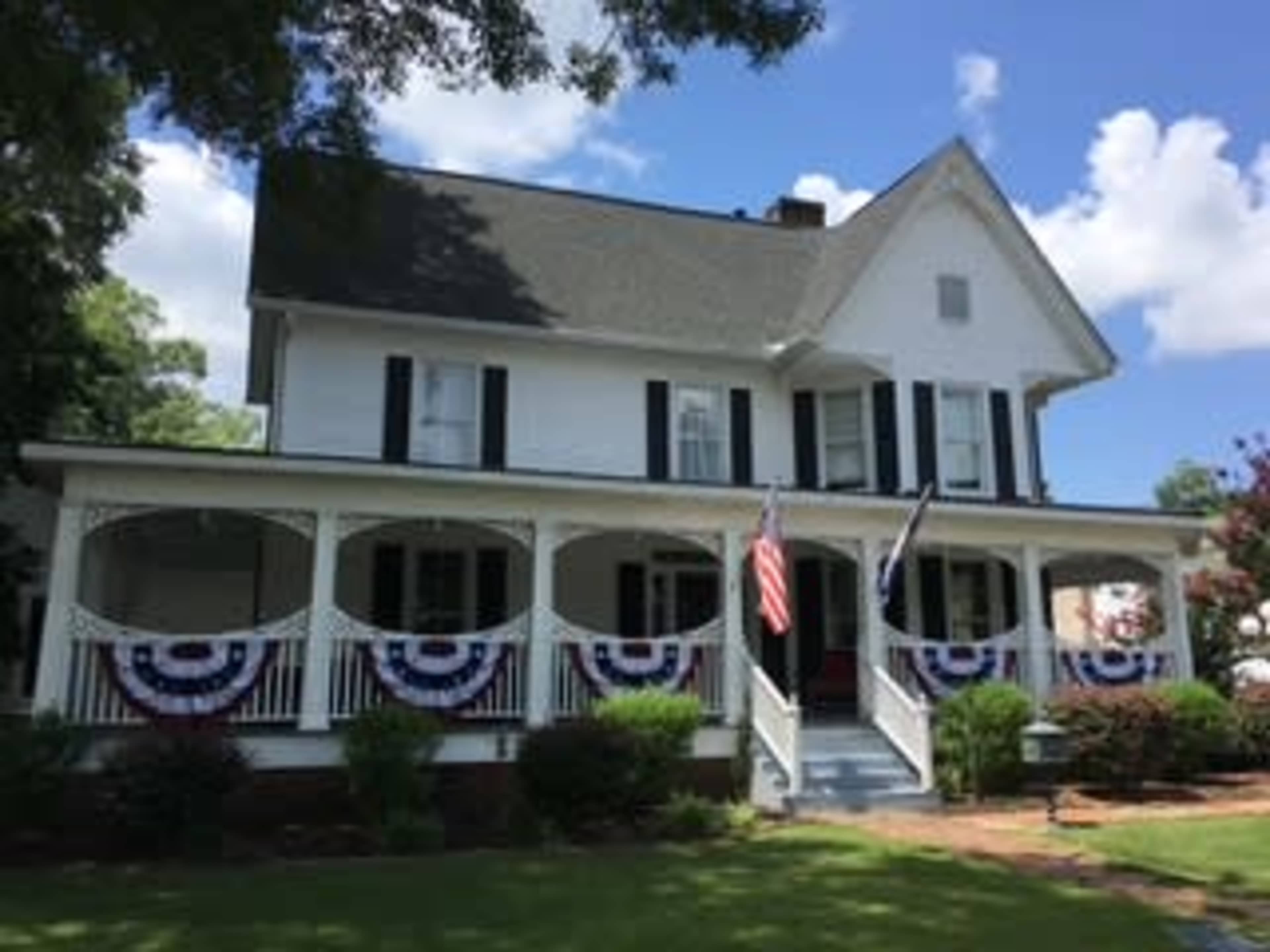 A two-story white house features a wraparound porch decorated with red, white, and blue bunting and American flags.