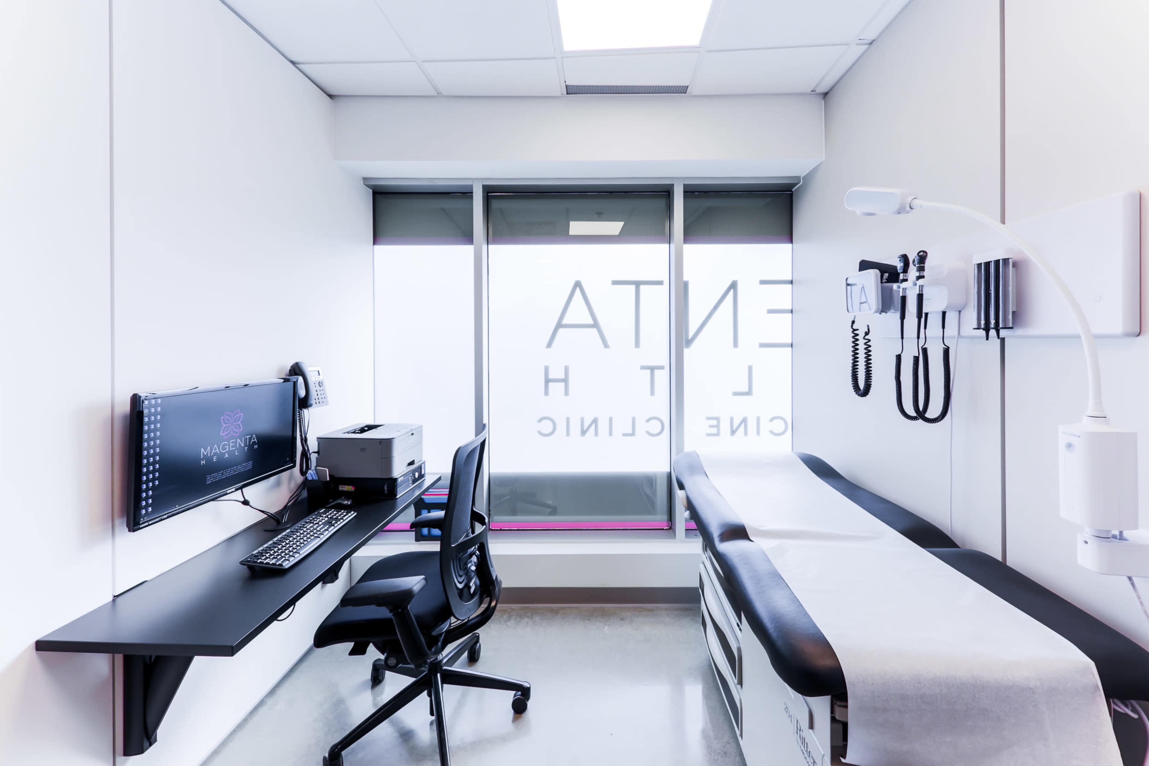 The image shows a modern medical examination room with a computer setup, a medical examination table, and medical equipment on the wall.