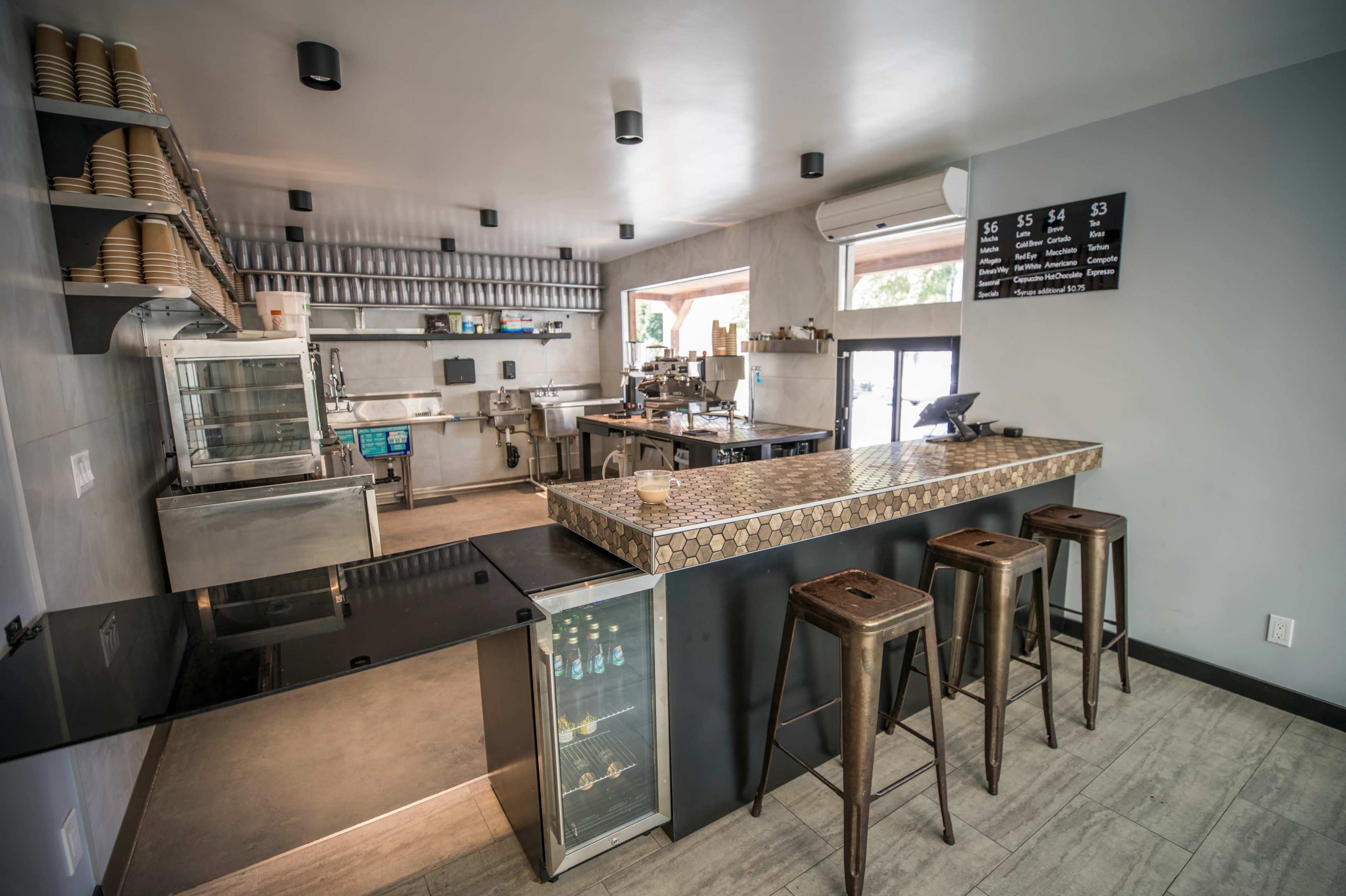A modern kitchen and bar area with metal stools, a countertop, and various kitchen equipment arranged along the walls.