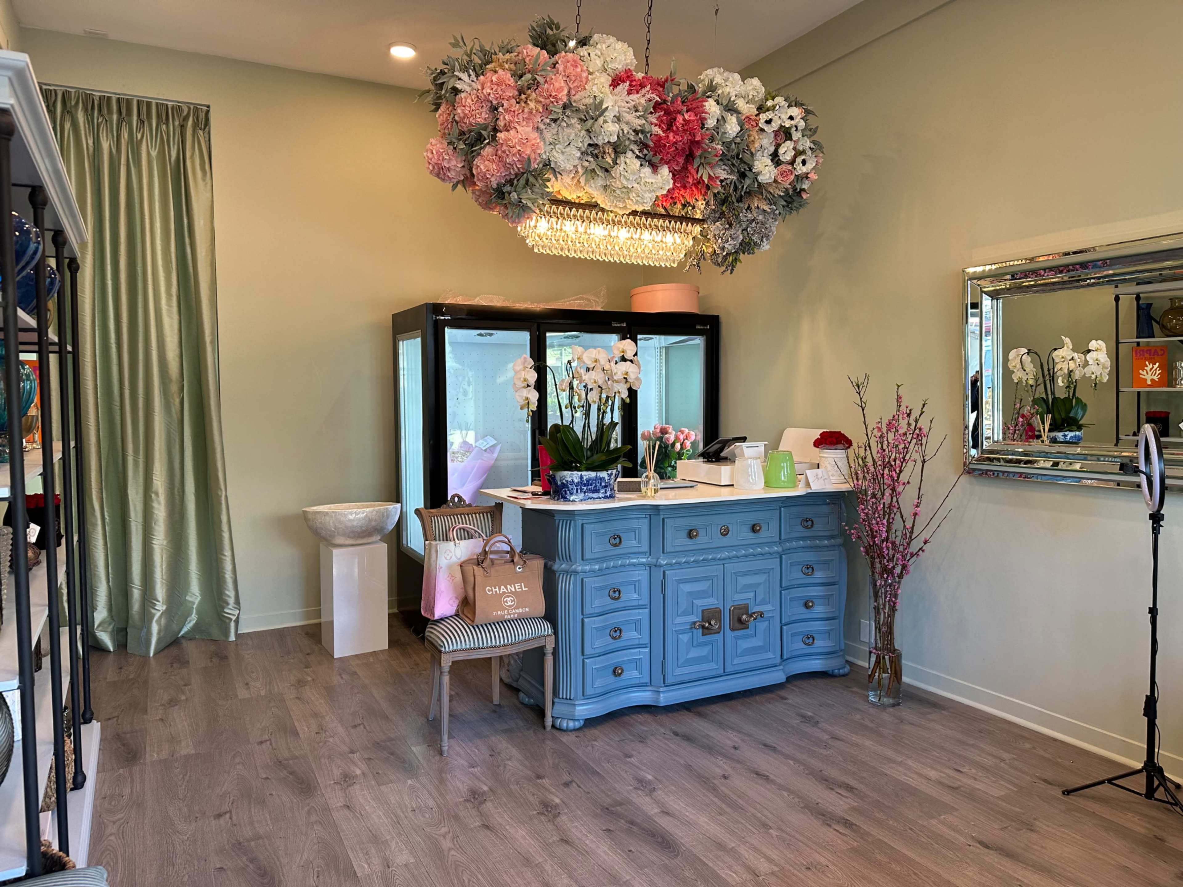 The image shows a boutique interior featuring a light blue dresser, a large floral chandelier, glass display cases, and decorative elements like potted orchids and a tall mirror.