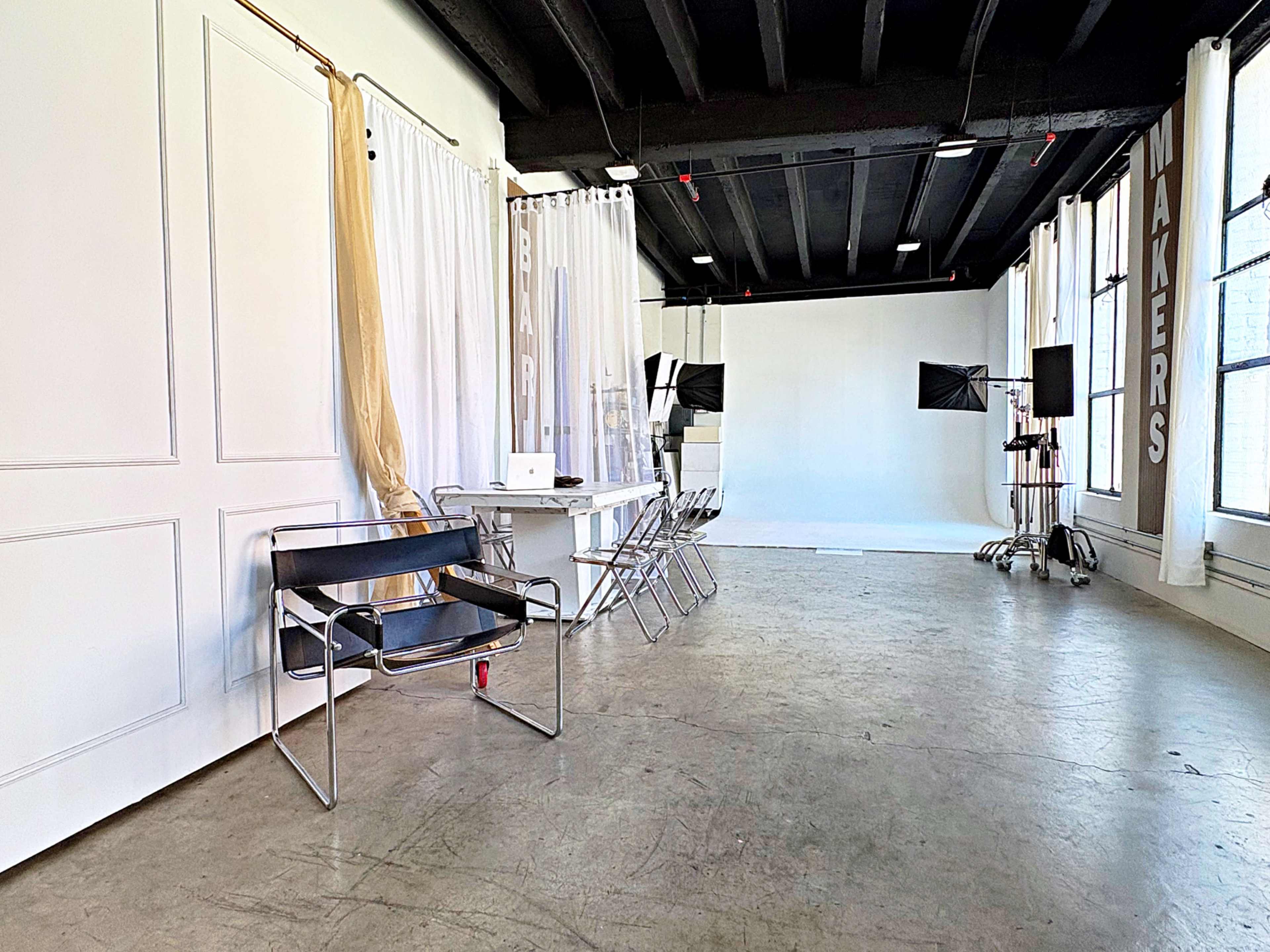The image shows a spacious studio with white walls and large windows, featuring a table with a computer, several chairs, and lighting equipment set against a plain backdrop.