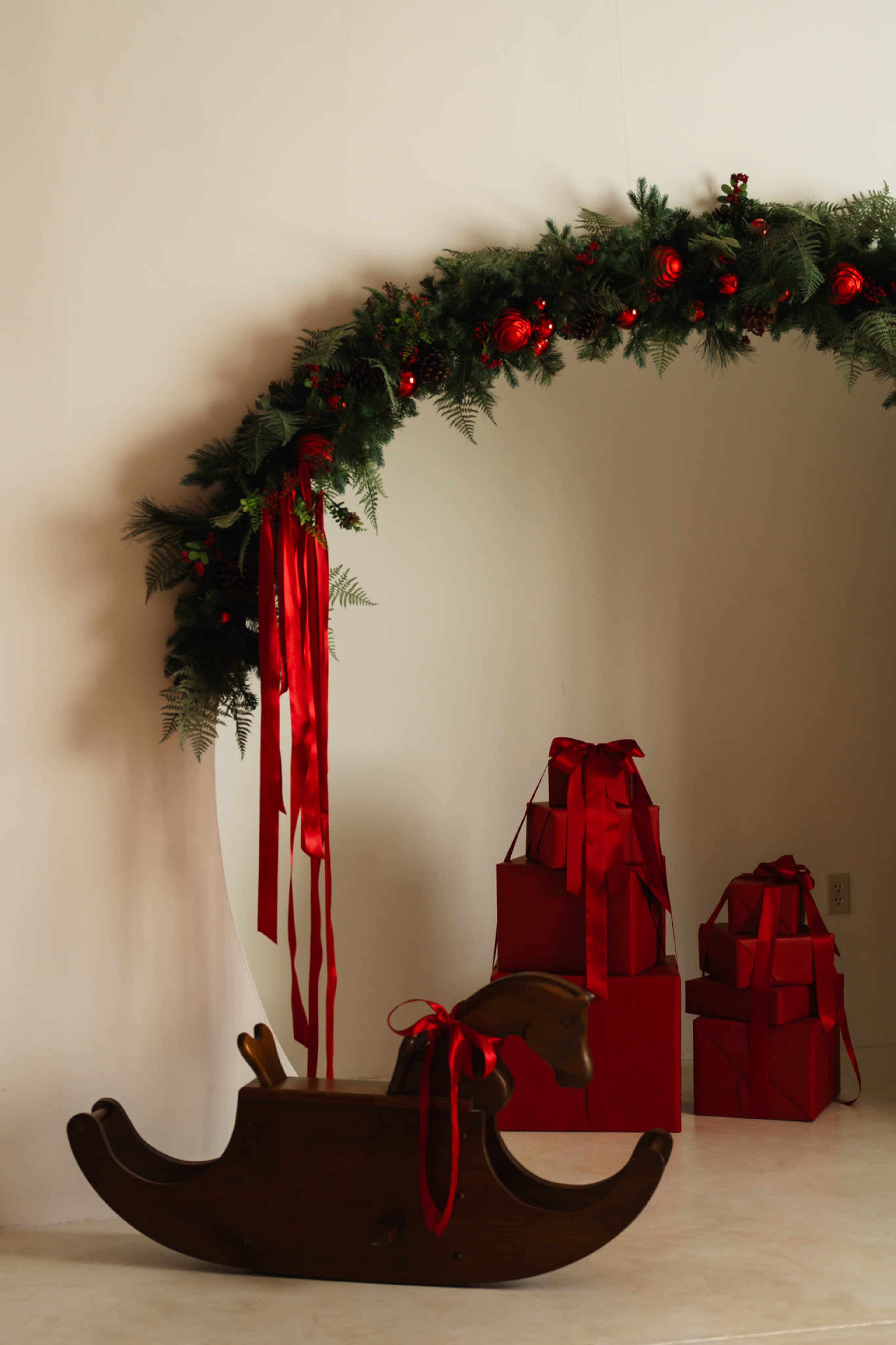 A decorated archway with greenery and red ornaments frames a wooden rocking horse and several red gift boxes.
