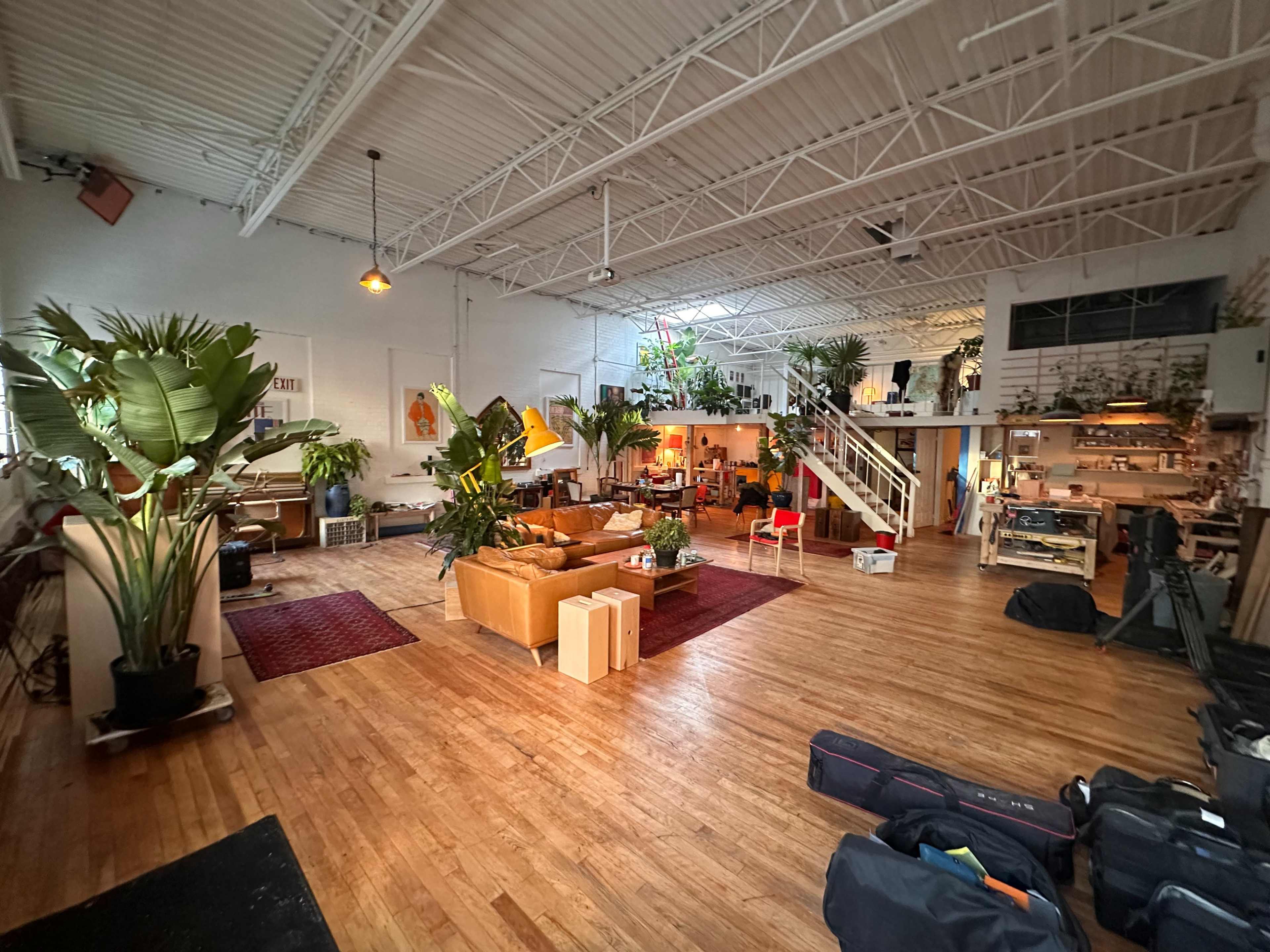 The image shows a spacious loft-style interior with wooden floors, a large seating area, potted plants, and various furniture arrangements.