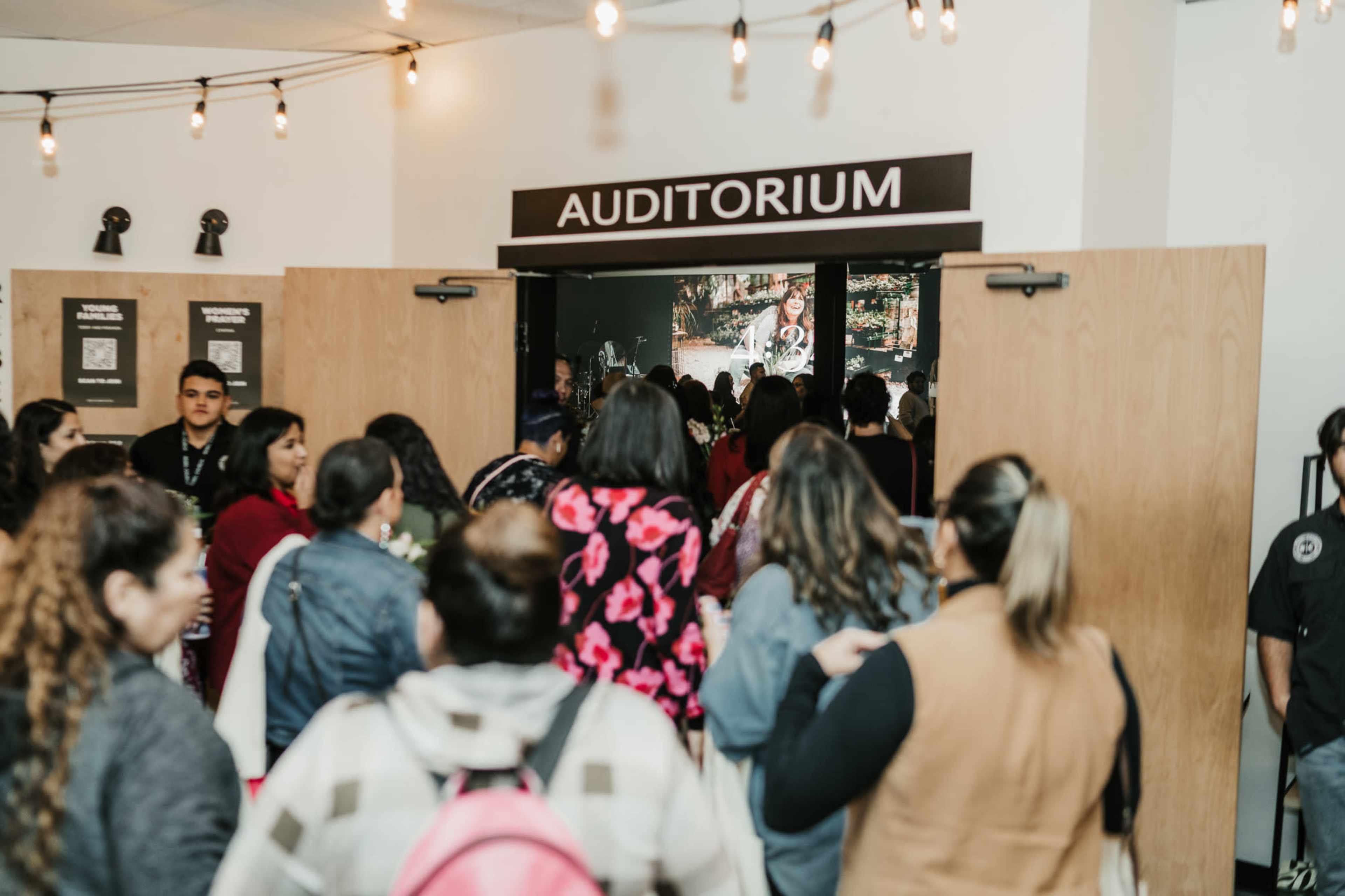A crowd of people gathers at the entrance of an auditorium, with the doors open and a screen visible inside.
