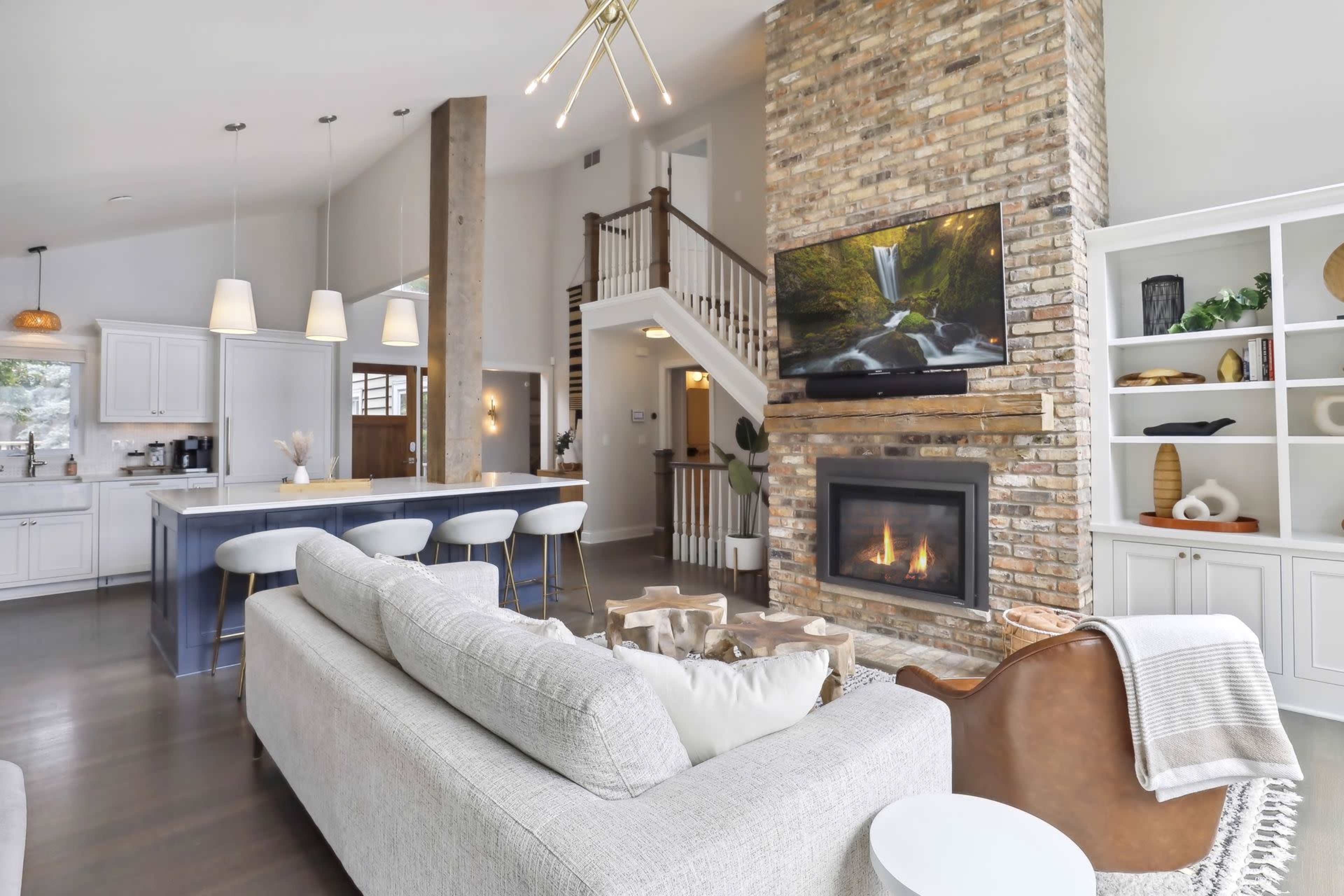 A modern living room with a stone fireplace, a large sectional sofa, and an adjacent kitchen featuring a central island with bar stools.