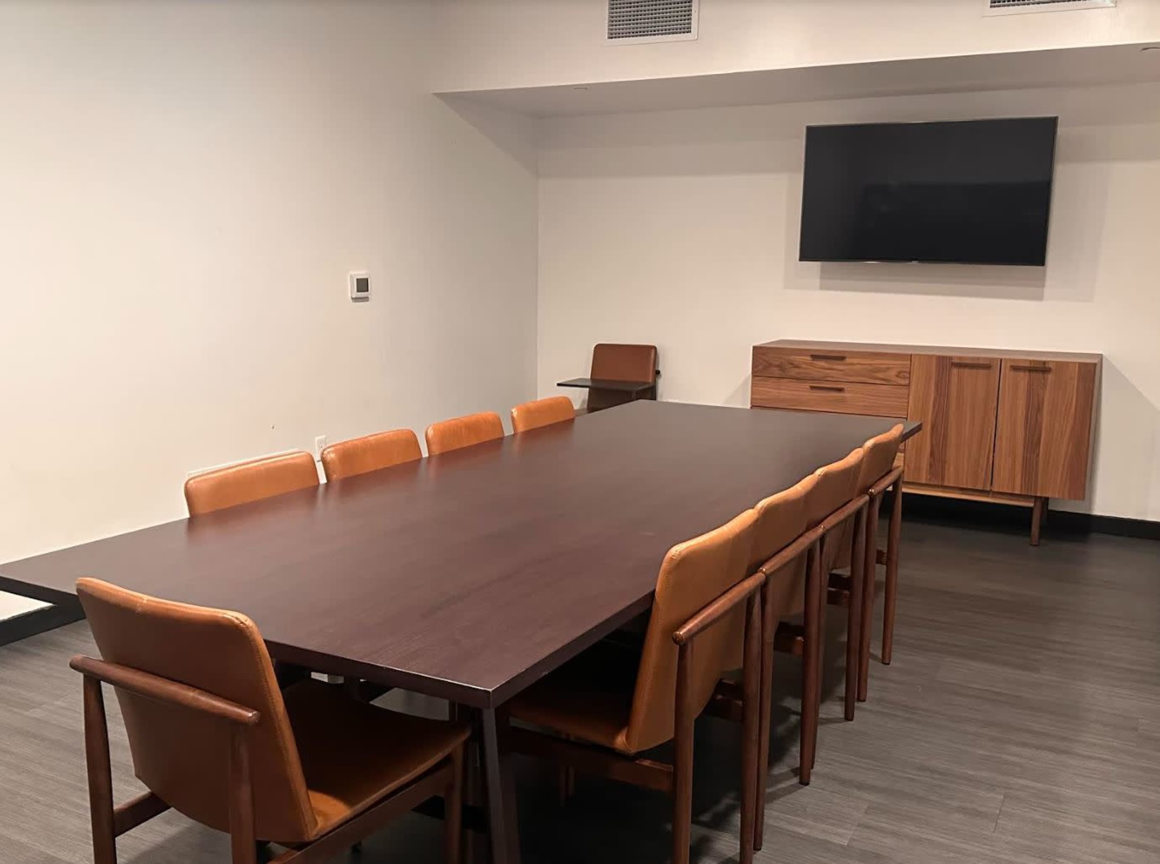 A large conference room features a long wooden table surrounded by eight brown chairs, with a television mounted on the wall and a side cabinet nearby.