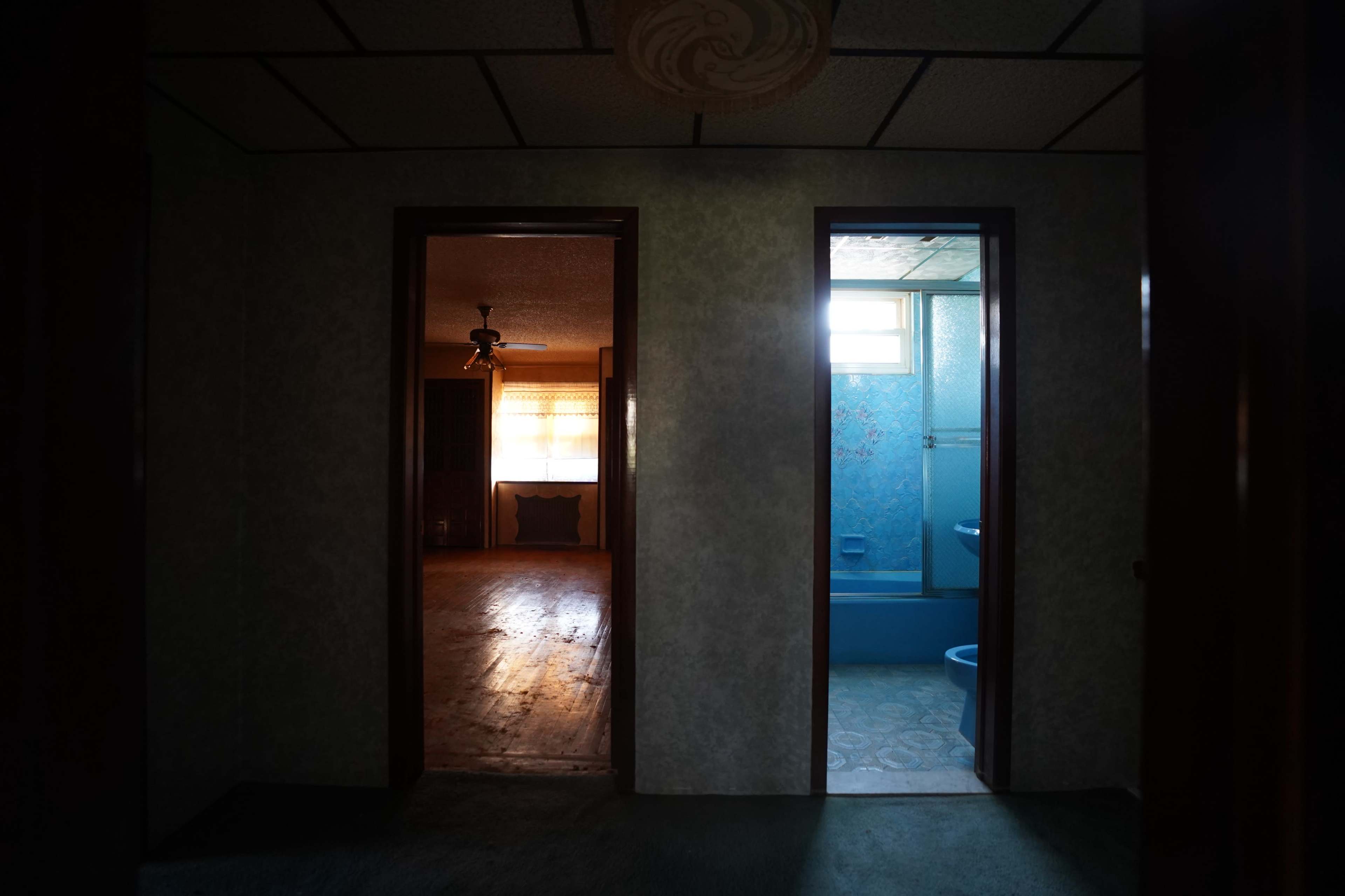 A dimly lit hallway separates two rooms, one with a visible window and wooden floor, and the other leading to a blue-tiled bathroom.