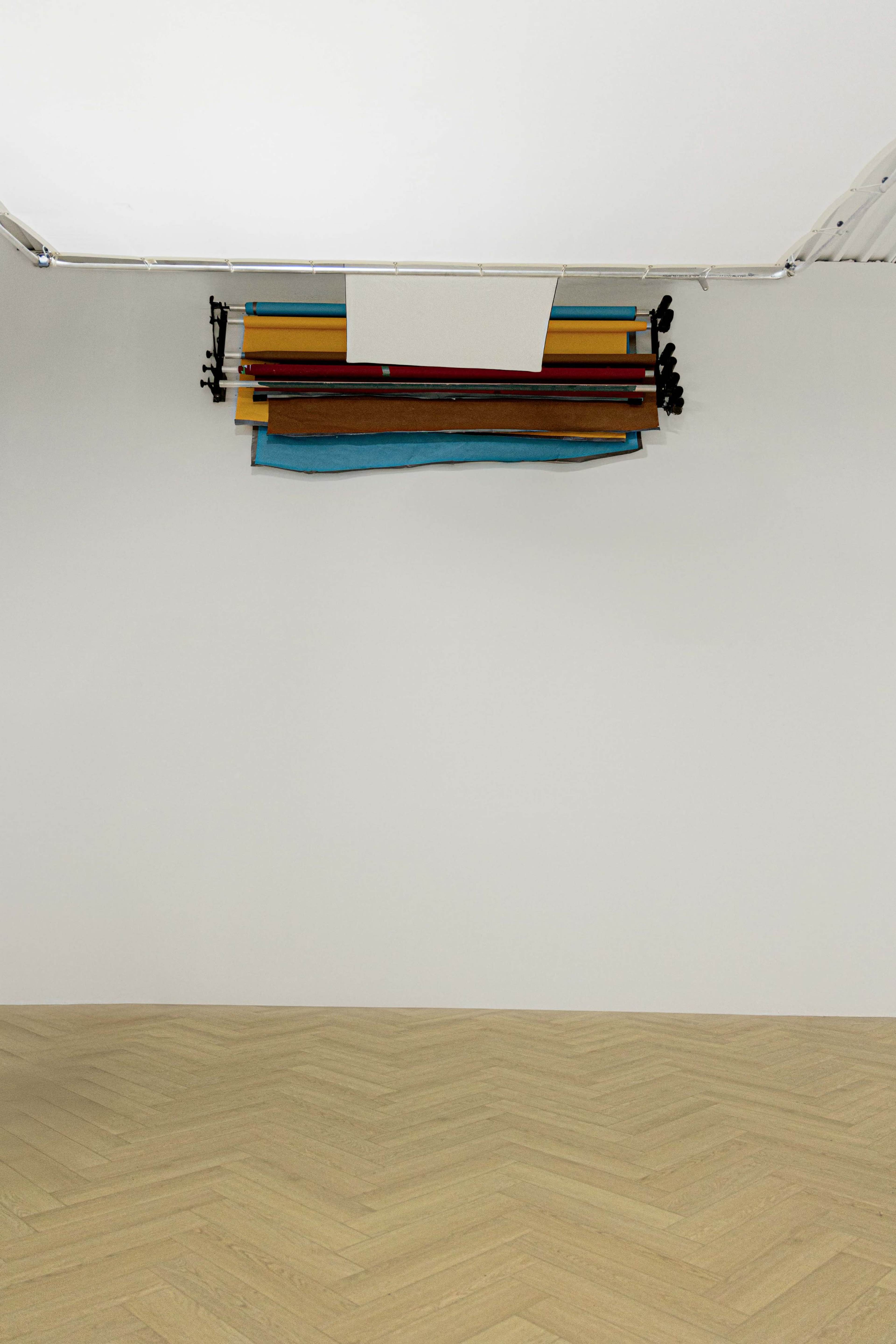 The image shows a minimalist interior space with a light wooden floor and a collection of colorful materials suspended from the ceiling against a plain white wall.