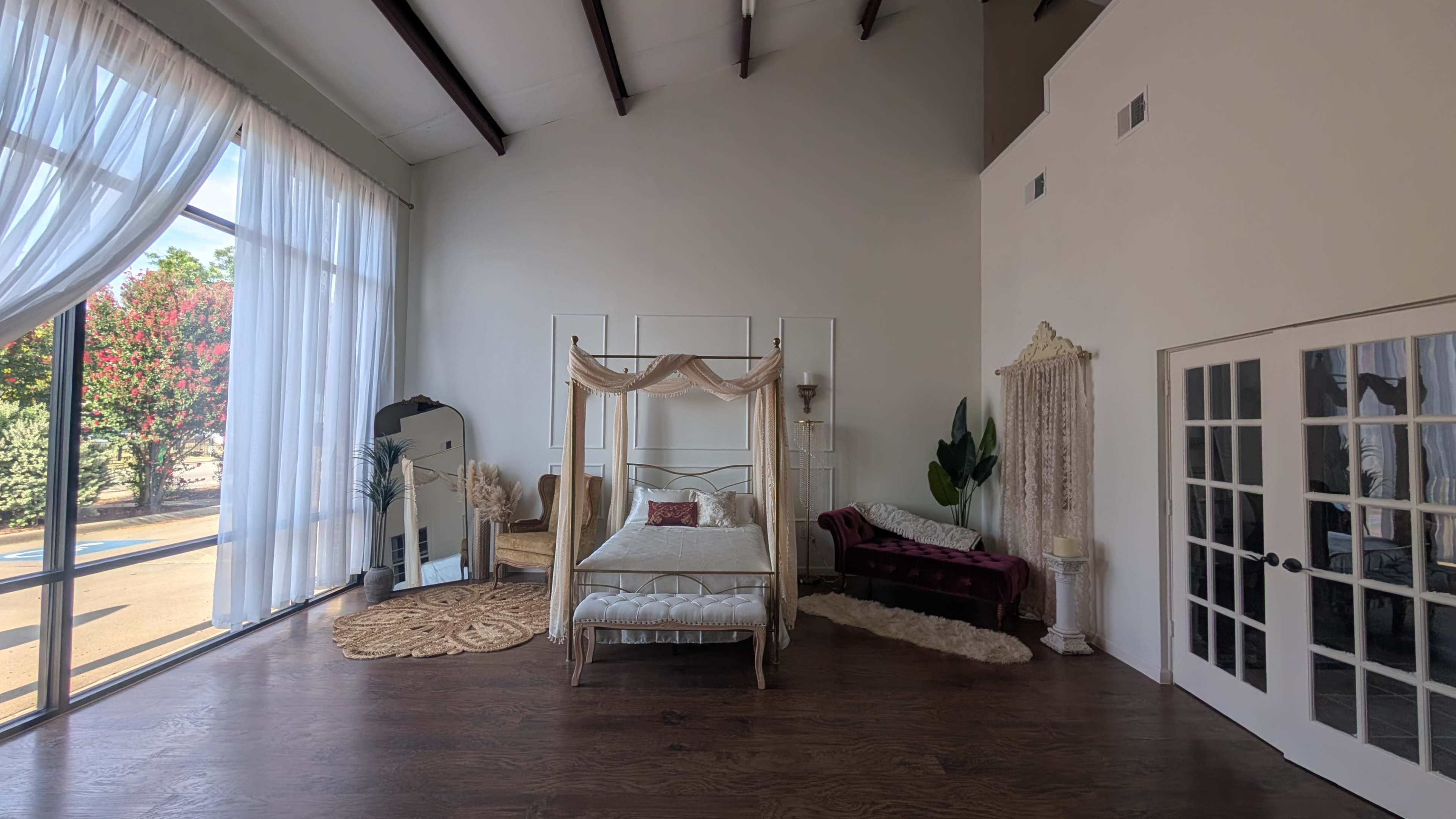 The image shows a spacious, light-filled room featuring a four-poster bed, a velvet sofa, a large mirror, and a set of French doors.