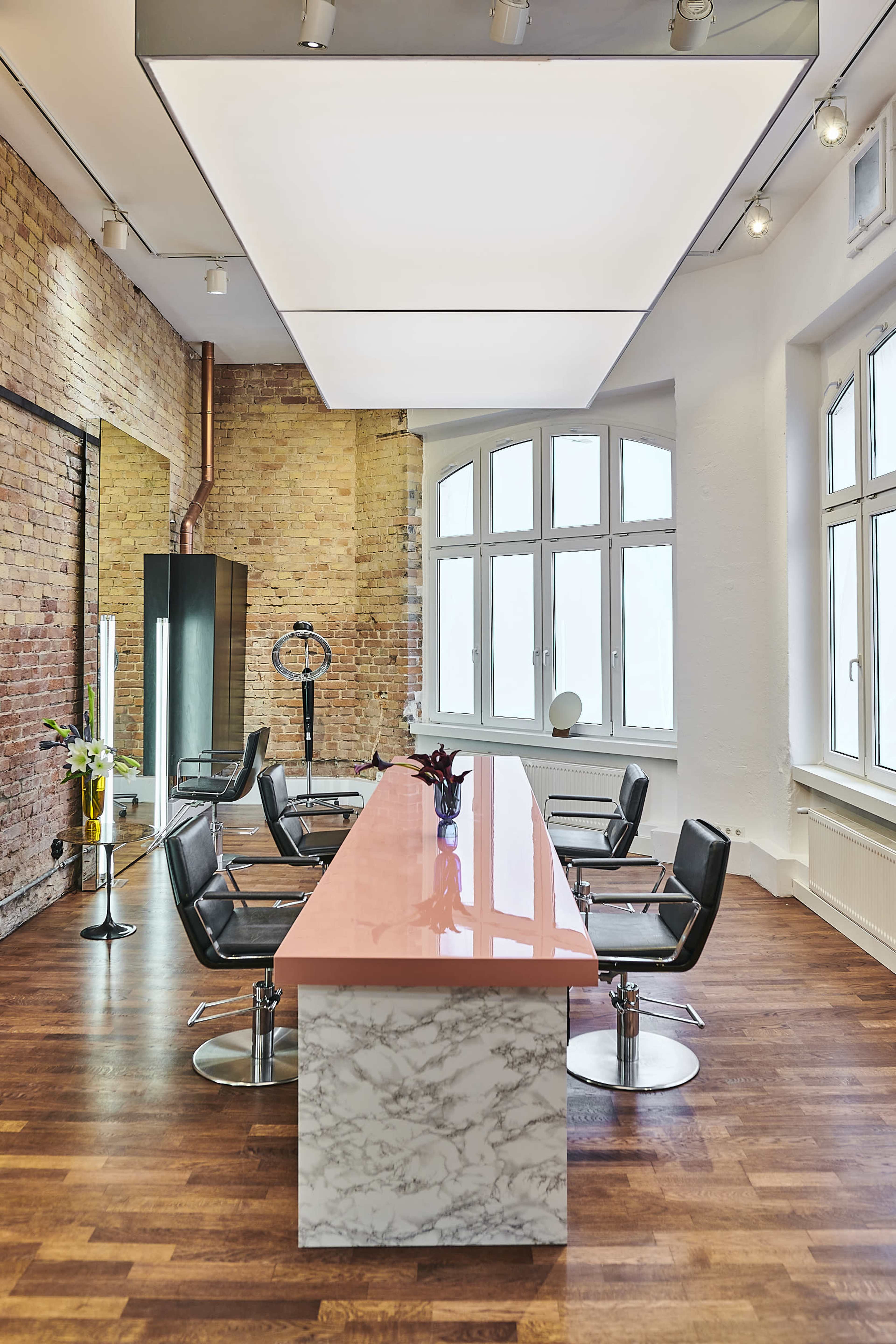 A modern conference room featuring a long pink marble table surrounded by black leather chairs, with large windows and brick walls in the background.