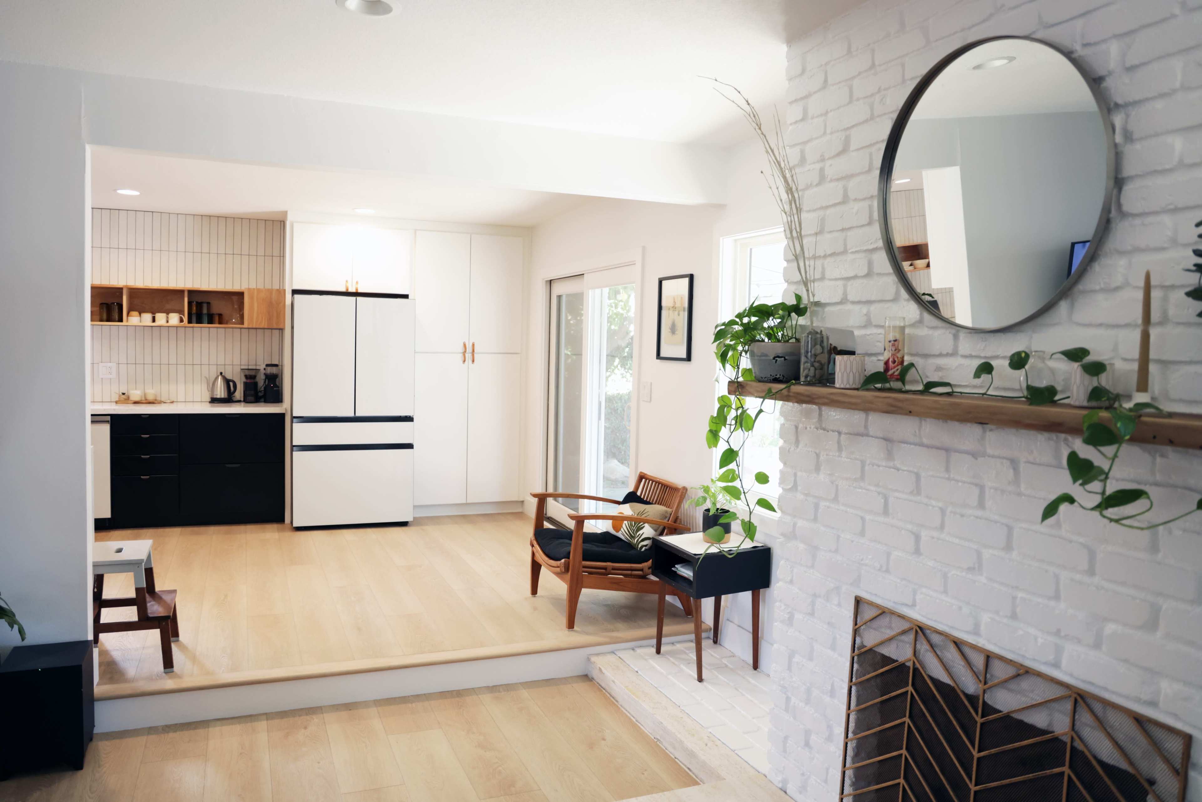 The image shows a modern living space featuring a kitchen area with black and white cabinetry, a wooden floor, a circular mirror above a white brick mantel, and a seating area with plants.