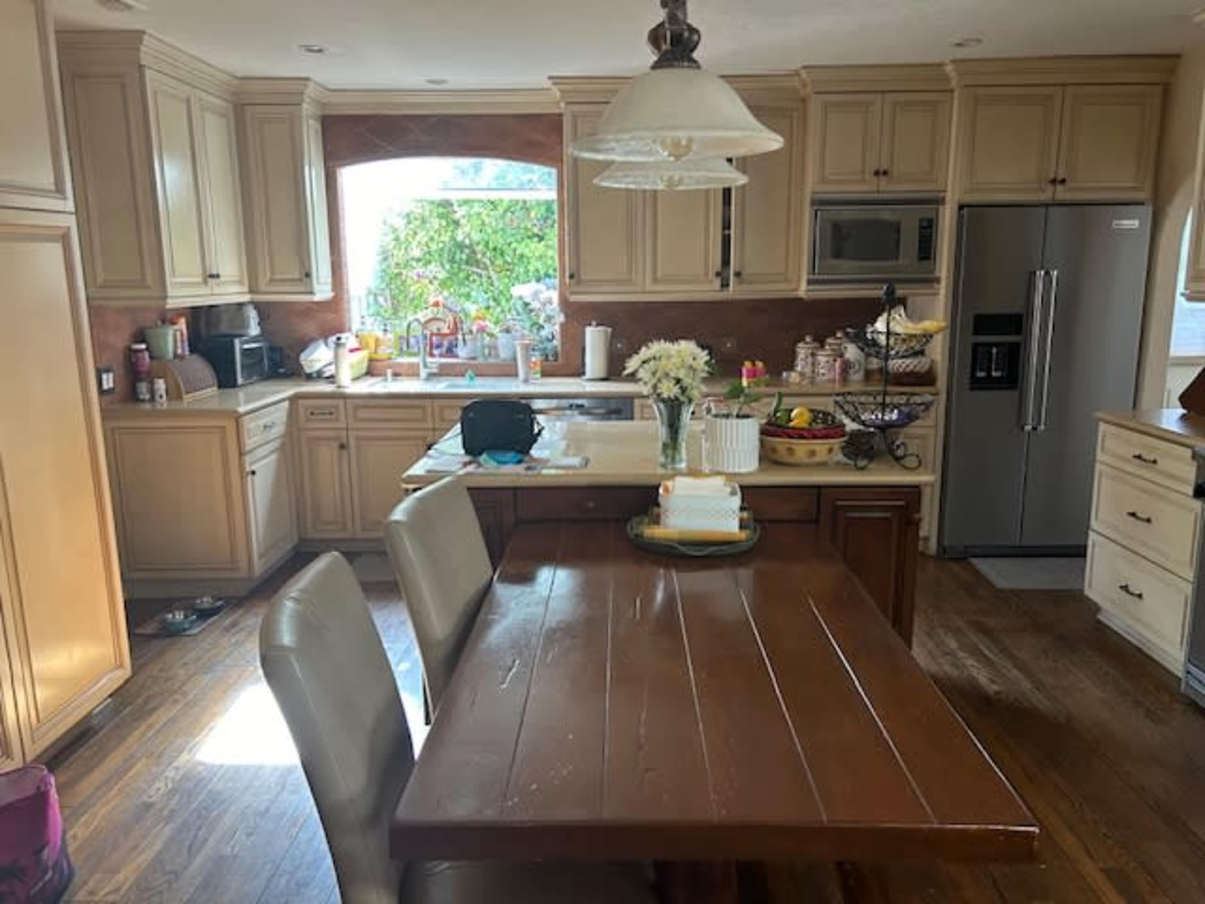 A well-lit kitchen with wooden cabinets, a central wooden dining table, and various appliances and items on the countertops.
