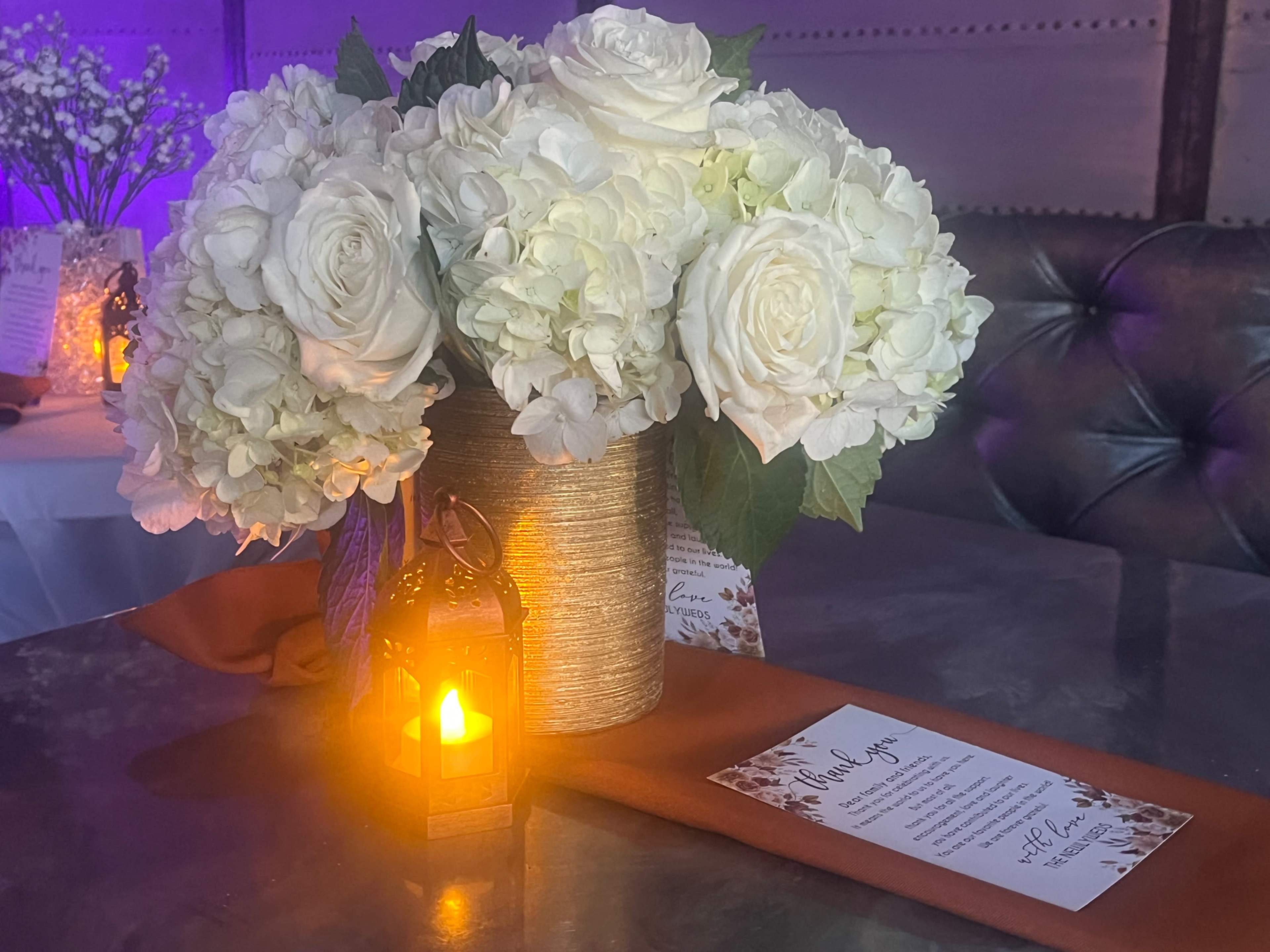 A floral arrangement of white roses and hydrangeas is displayed in a silver vase next to a small lantern and a menu on a table.