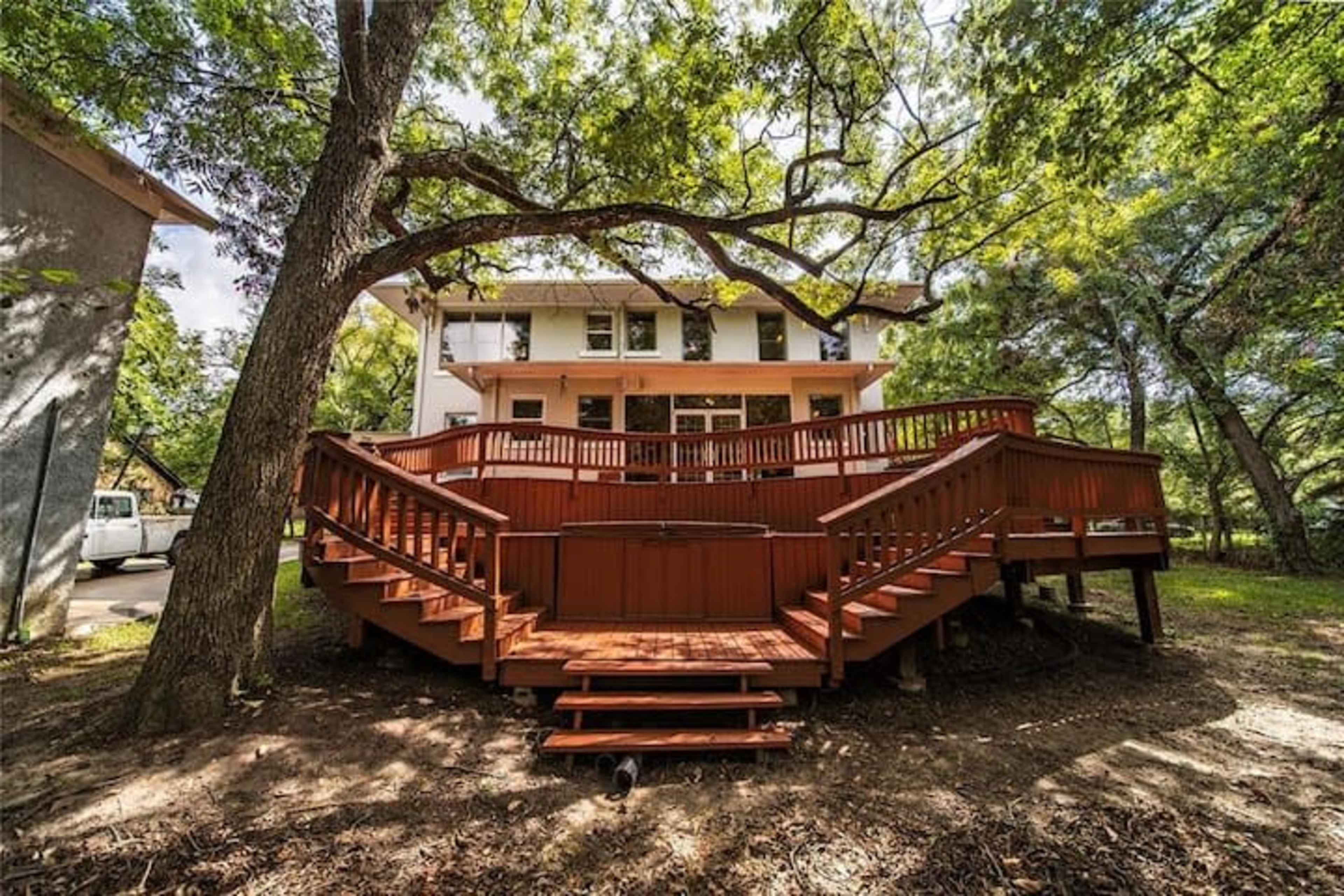 A large wooden deck surrounds the back of a two-story house, set among several trees in a grassy area.