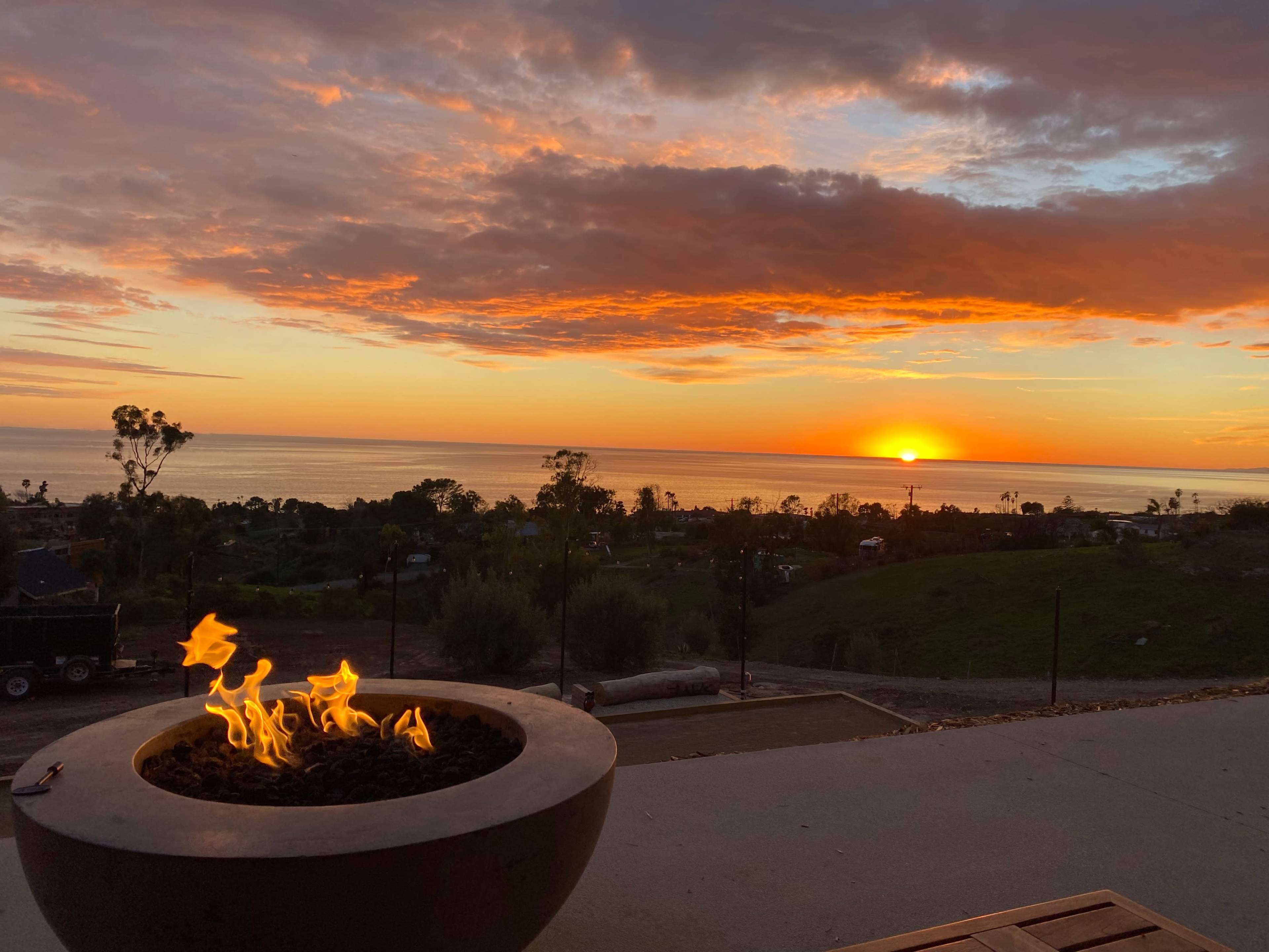 A fire pit with flames is positioned in the foreground while a sunset illuminates the ocean and sky in the background.