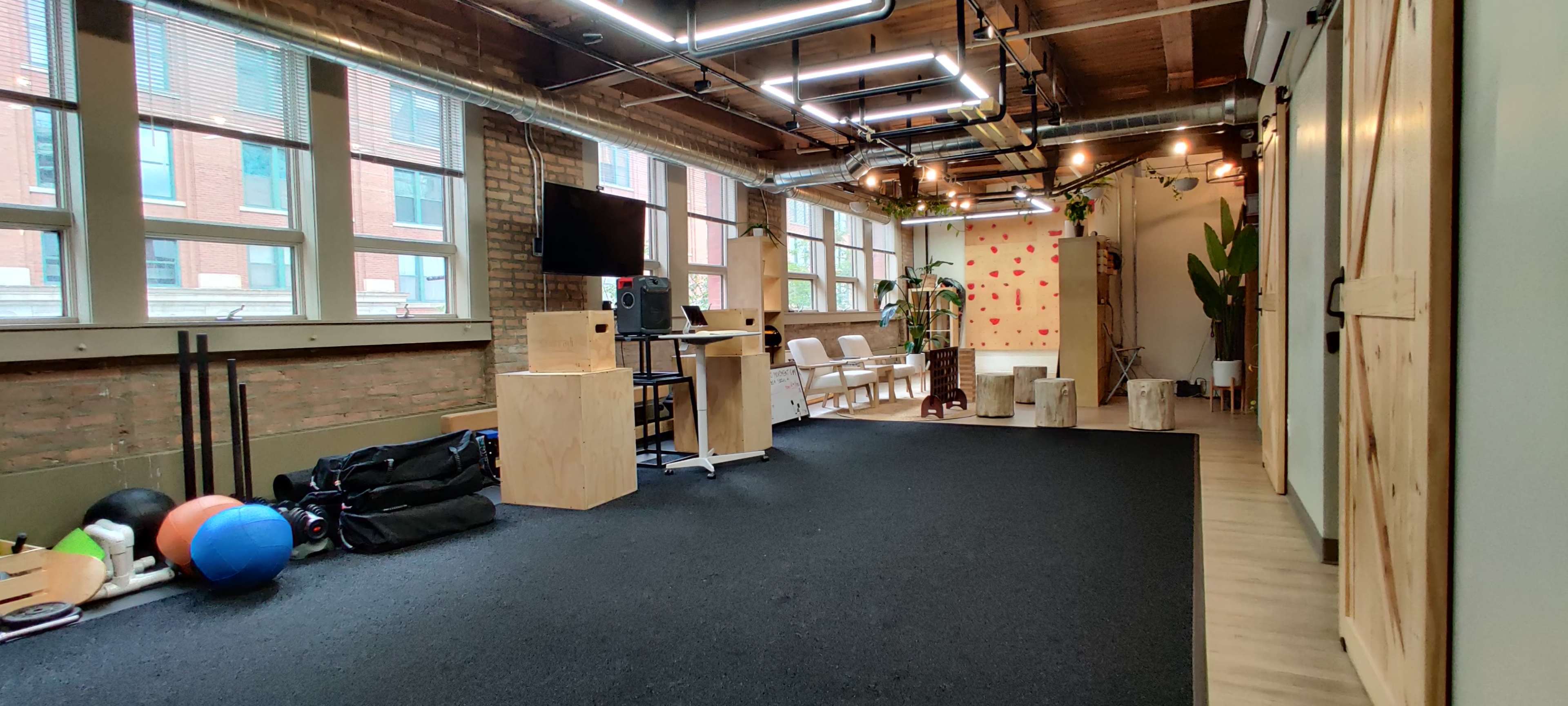 A spacious room with a black floor mat, large windows, wooden accents, workout equipment, and seating arrangements.