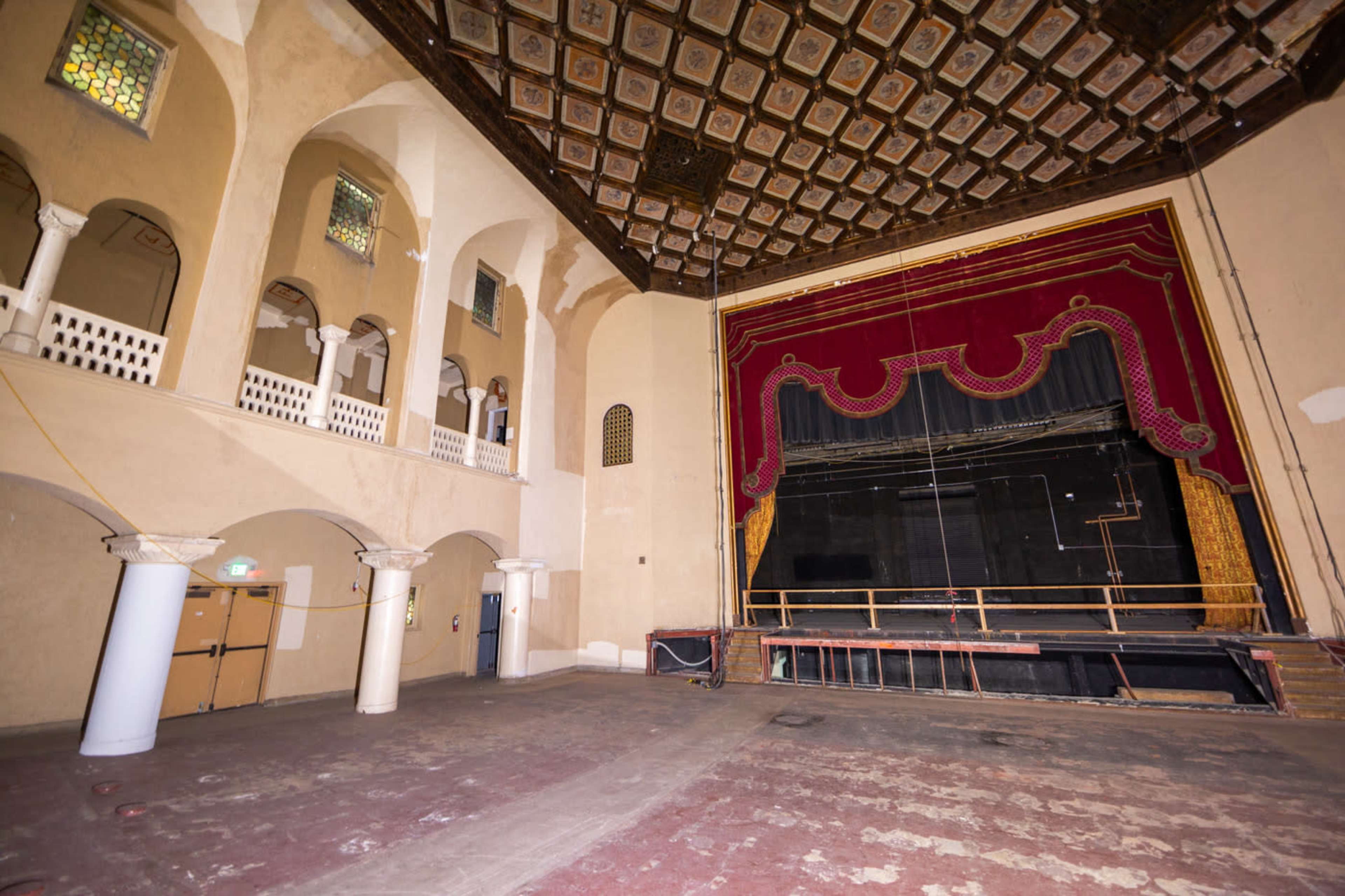 The image shows an empty theater with a proscenium stage, ornate ceiling, and arched windows.