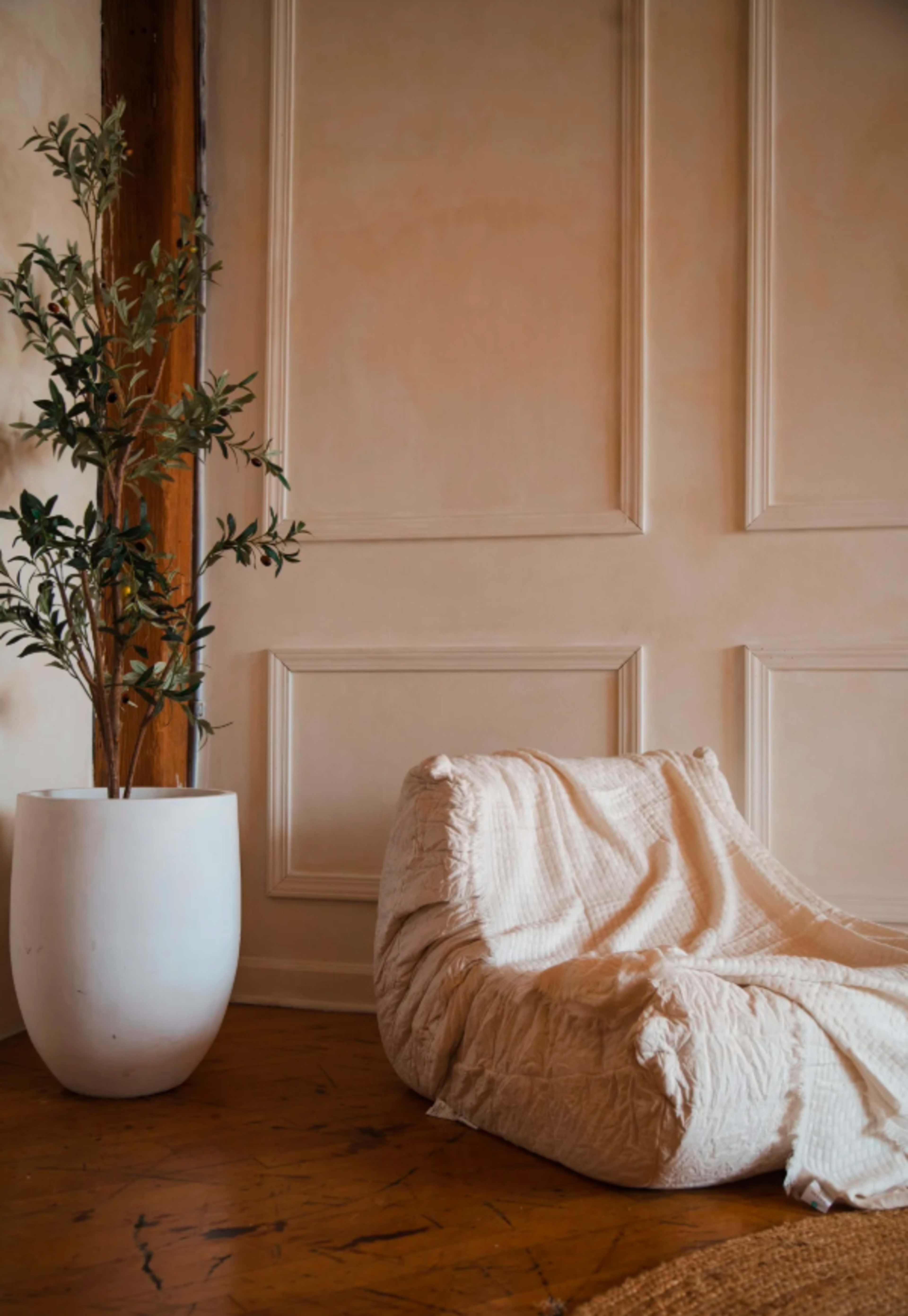 A white beanbag chair draped with a textured blanket sits next to a tall white planter containing leafy greenery against a plain wall.