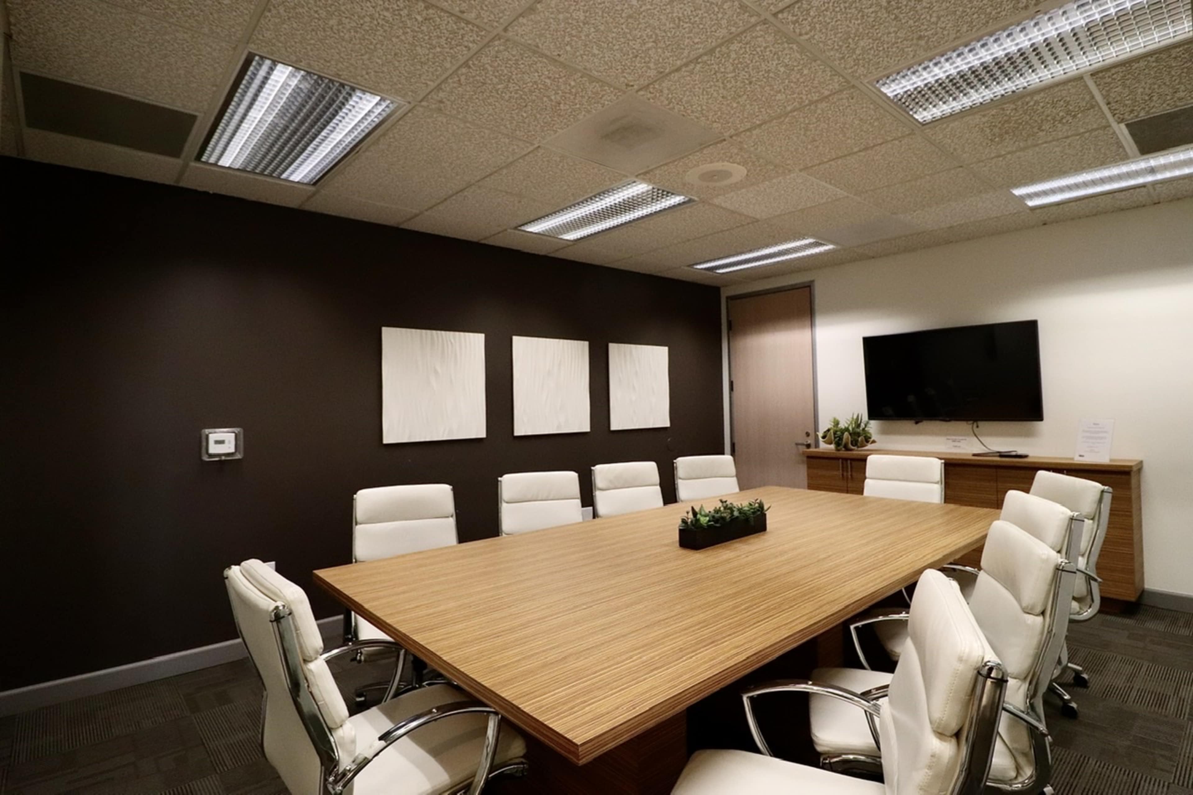 A modern conference room features a long wooden table surrounded by white leather chairs, with three framed artworks on the wall and a television mounted on the opposite wall.