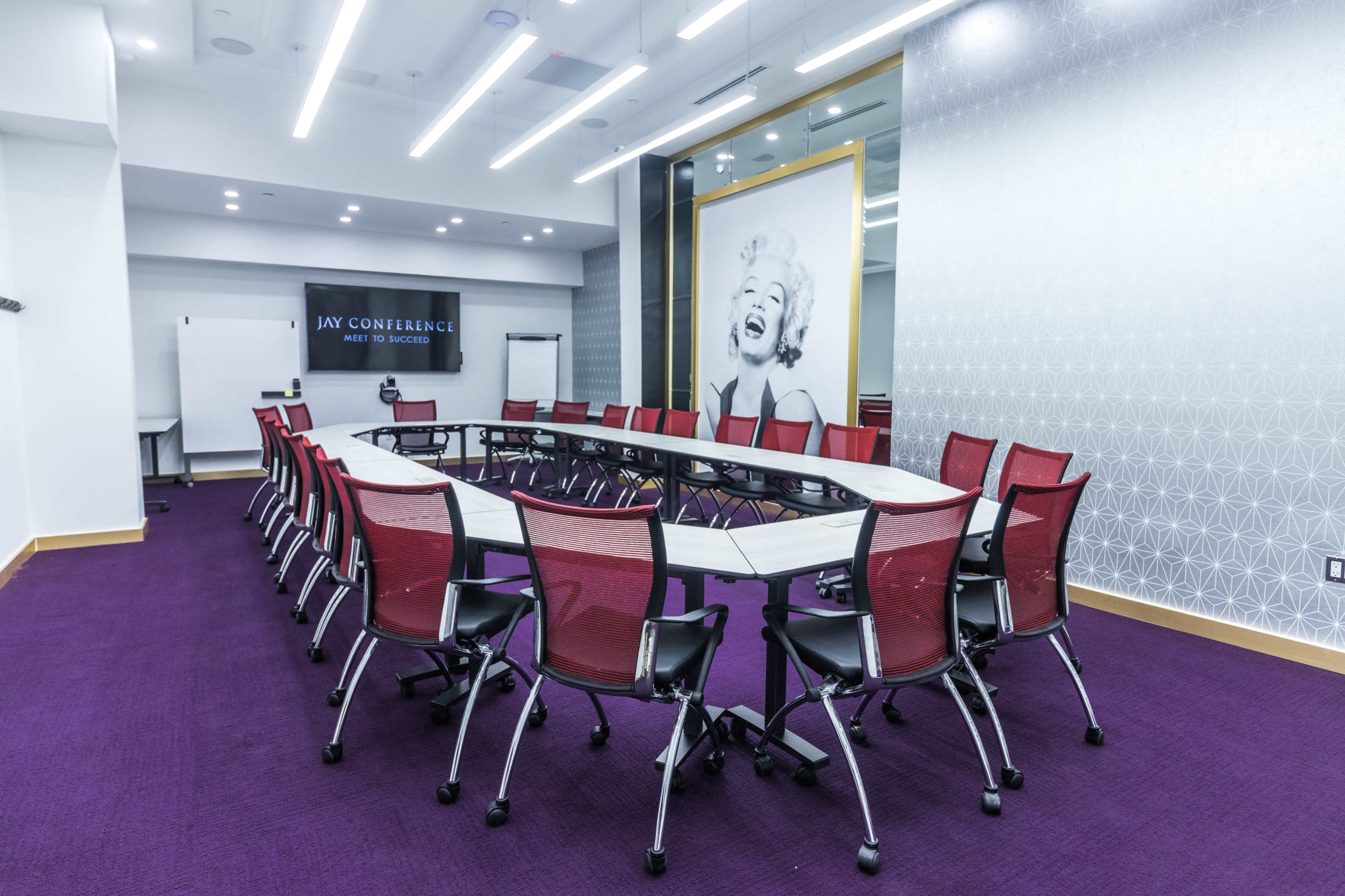 A modern conference room features a long, oval table surrounded by red mesh chairs, with a large image of a smiling woman on the wall.
