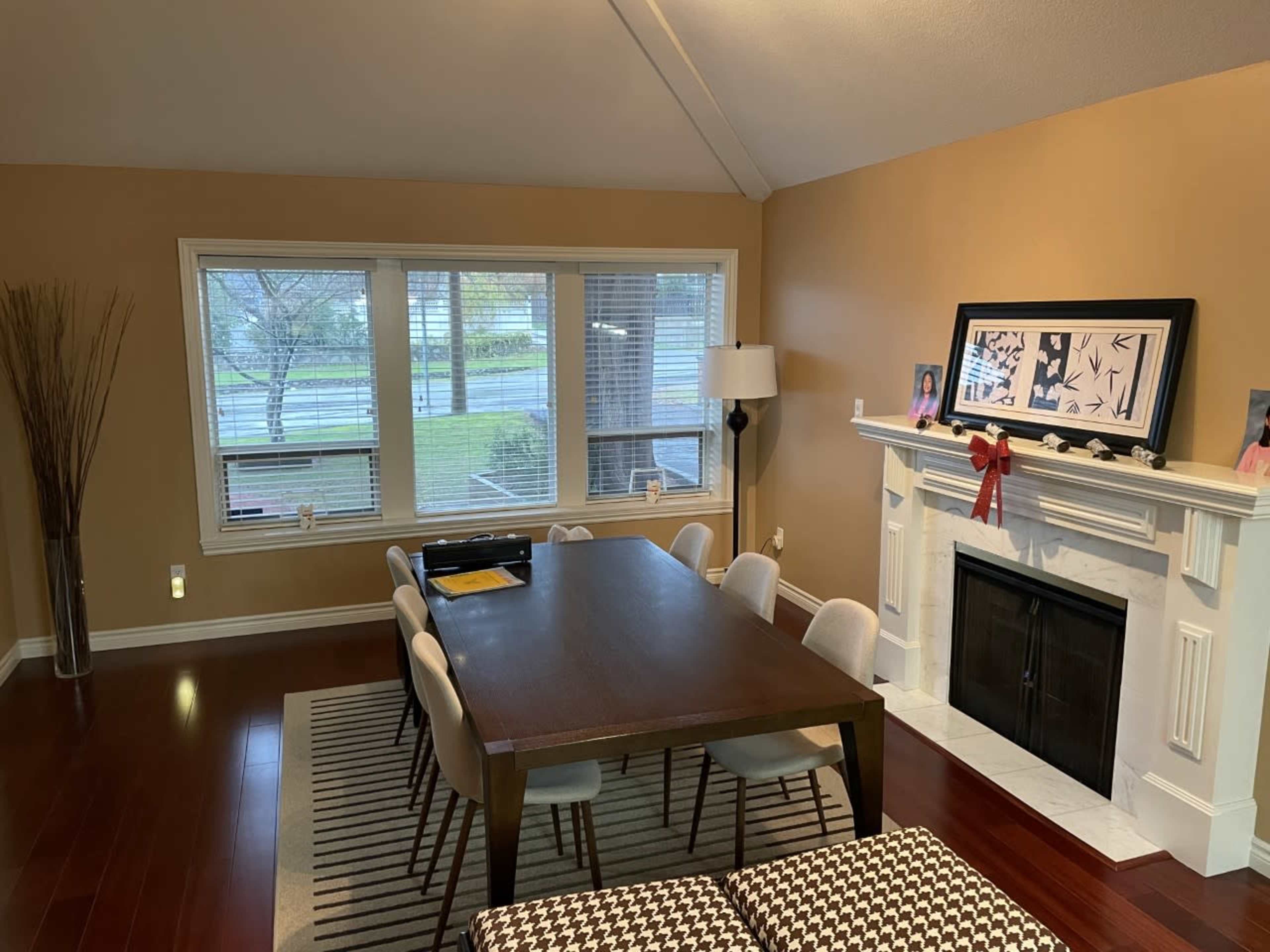 A dining room features a large wooden table surrounded by chairs, with a window offering a view of the outside and a fireplace with decorative elements.