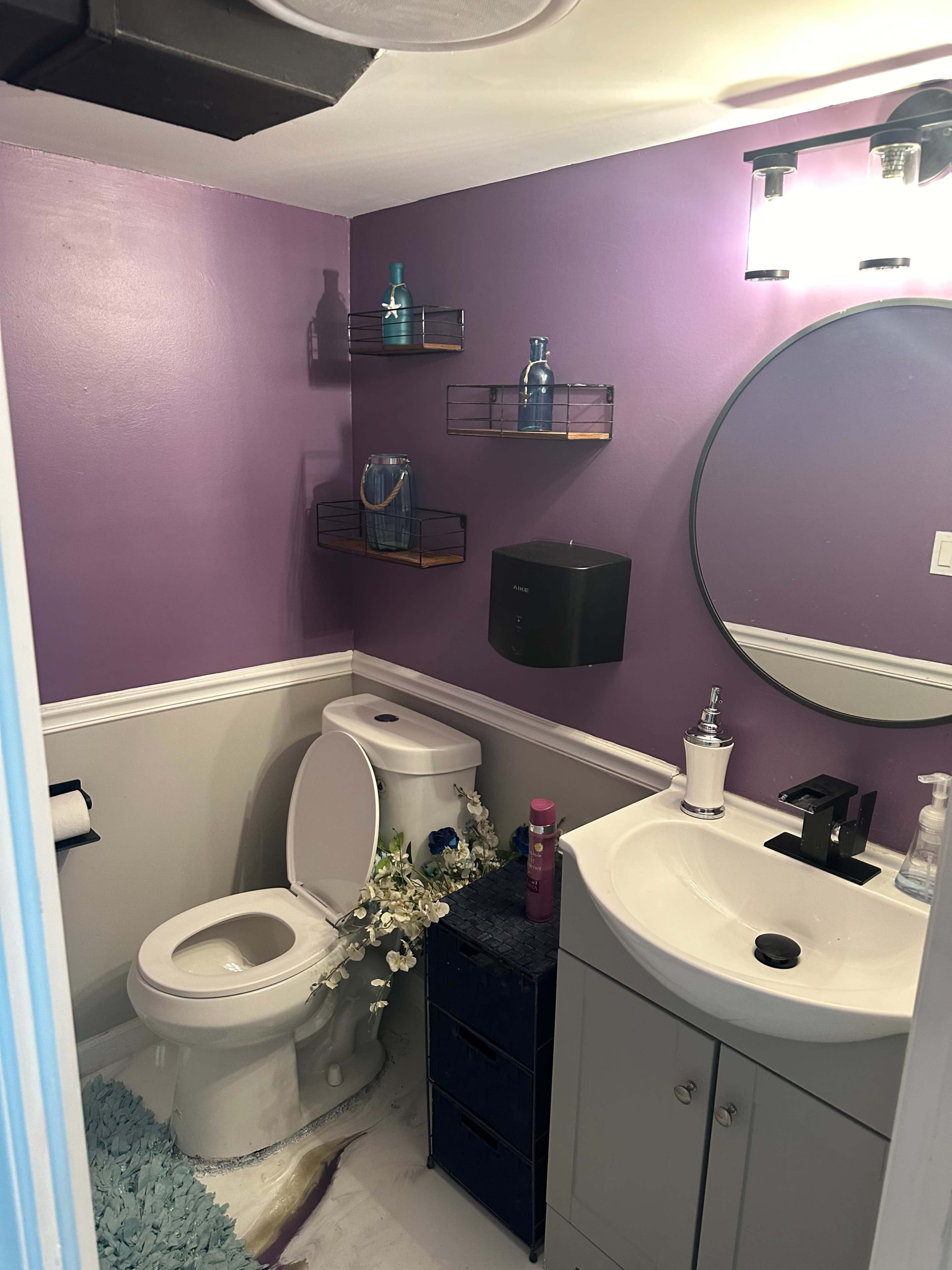 A small bathroom features a purple wall, a toilet, a sink with a dark cabinet, and decorative shelves with various toiletries.