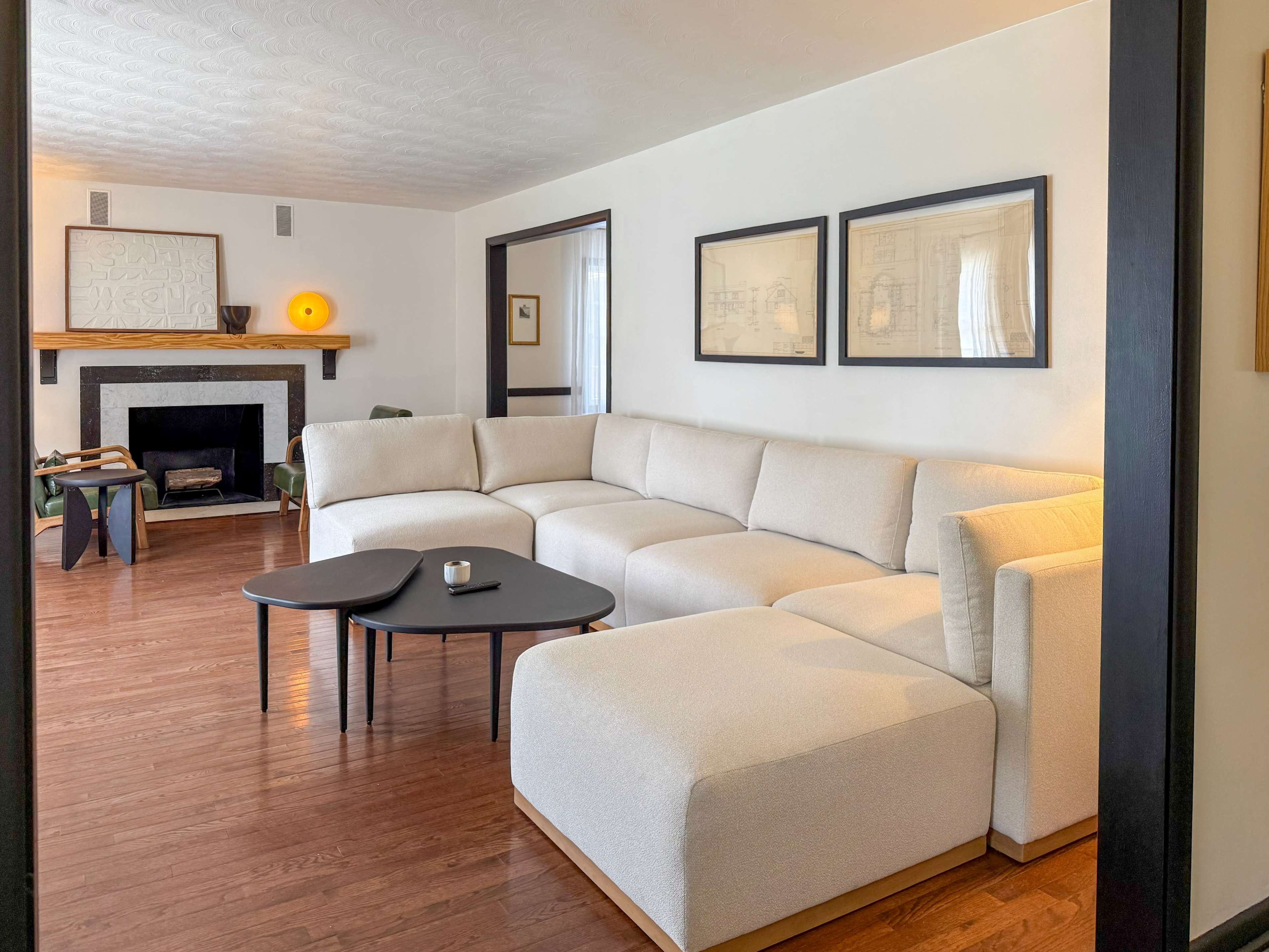 The image shows a modern living room with a large sectional sofa, a black coffee table, framed artwork on the walls, and a fireplace in the background.