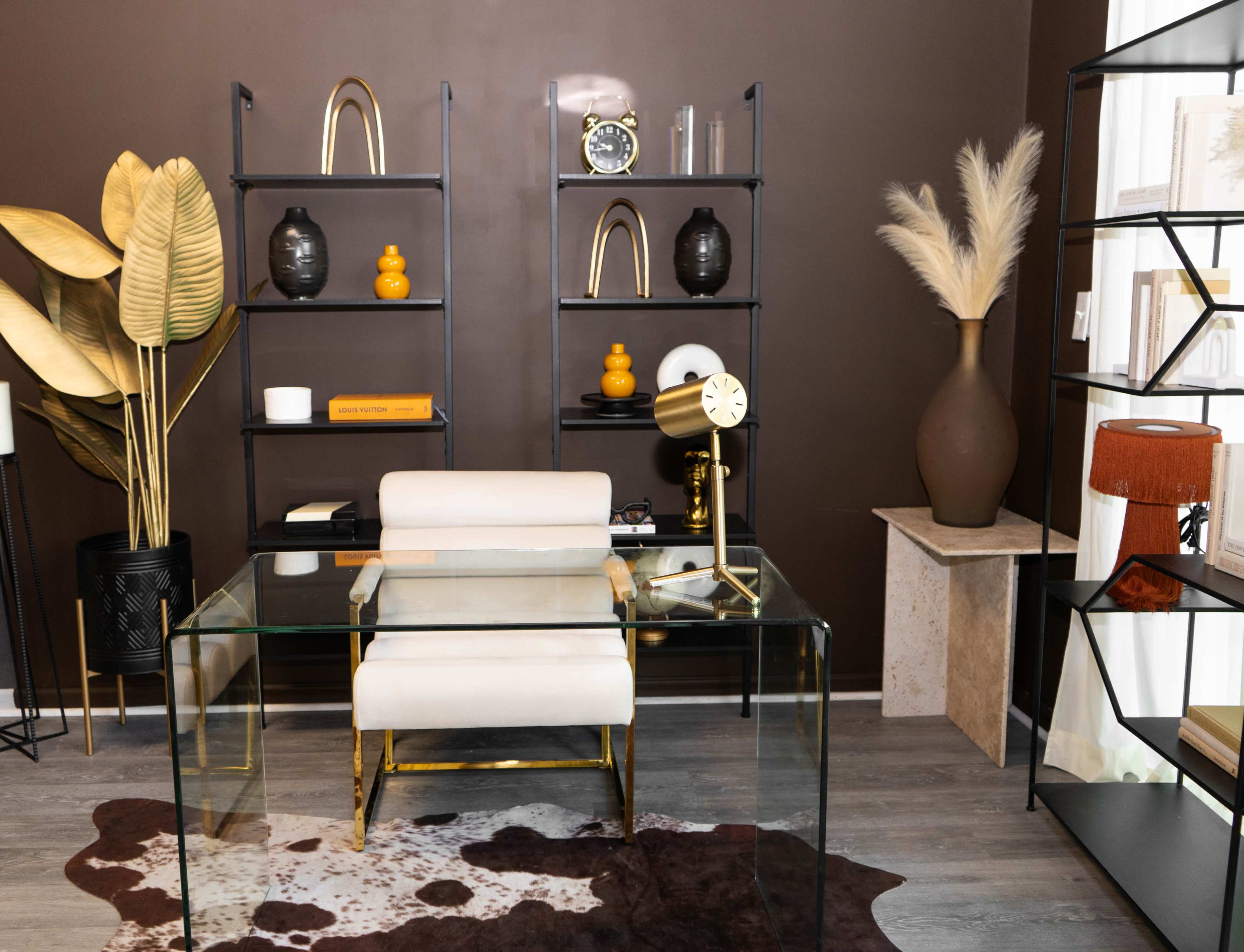 A modern office space featuring a glass desk, a white chair, and a variety of decorative items on black shelves against a brown wall.