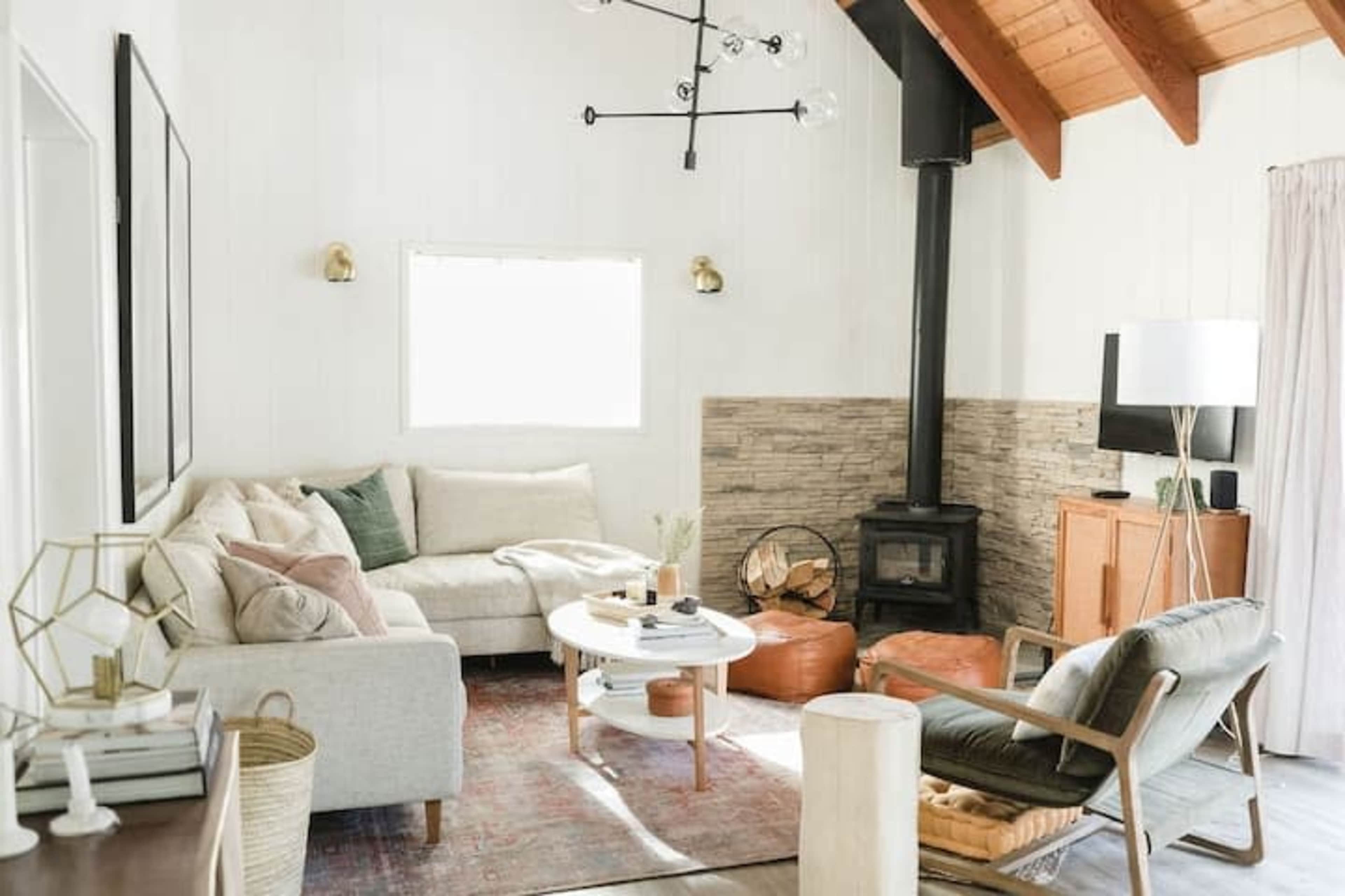 A living room features a light-colored sofa, a round coffee table, a wood stove, and a combination of wooden and stone elements.