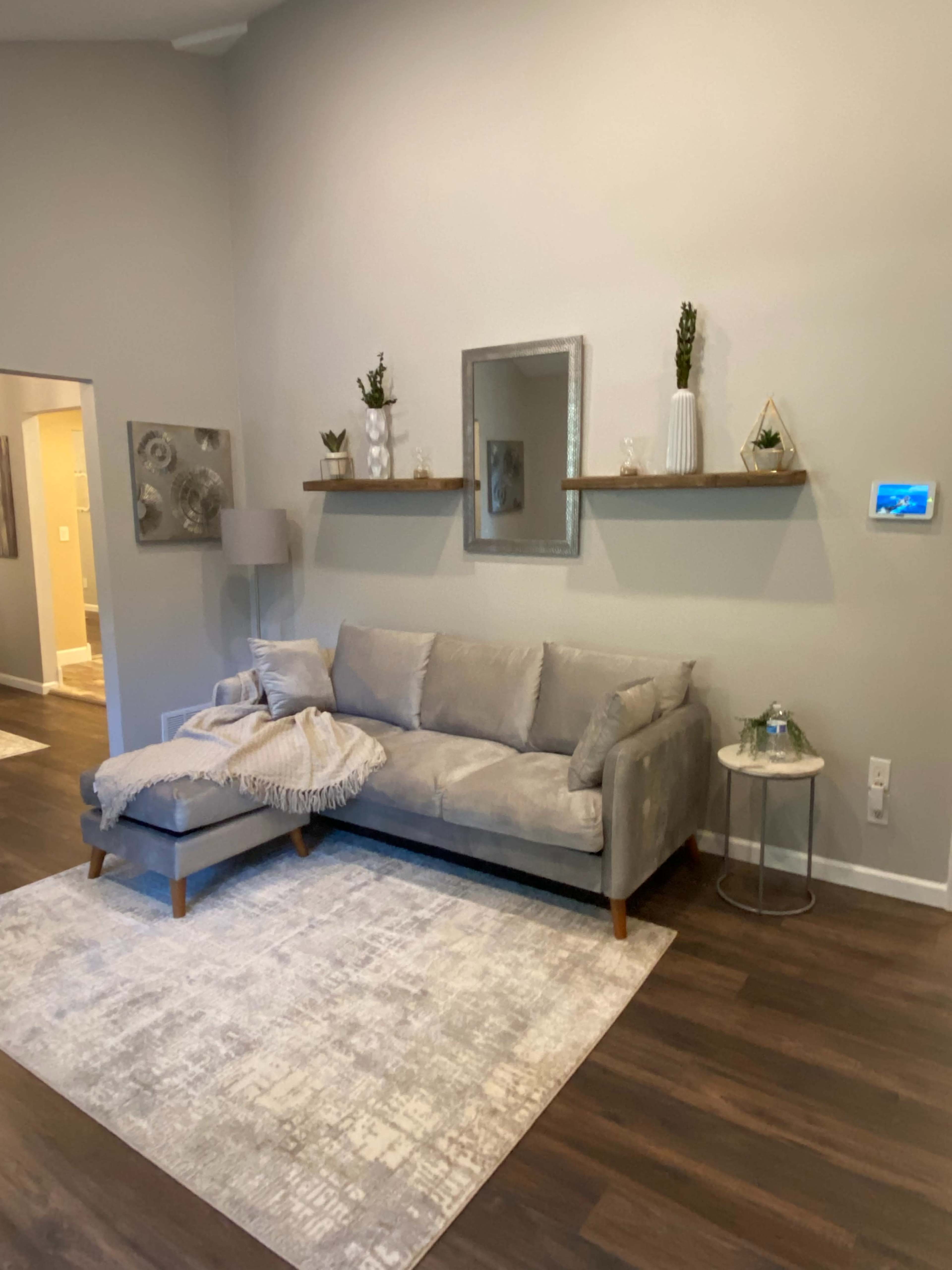A light gray couch with a throw and a small side table is positioned on a patterned rug against a neutral wall, featuring a mirror and decorative shelf above it.