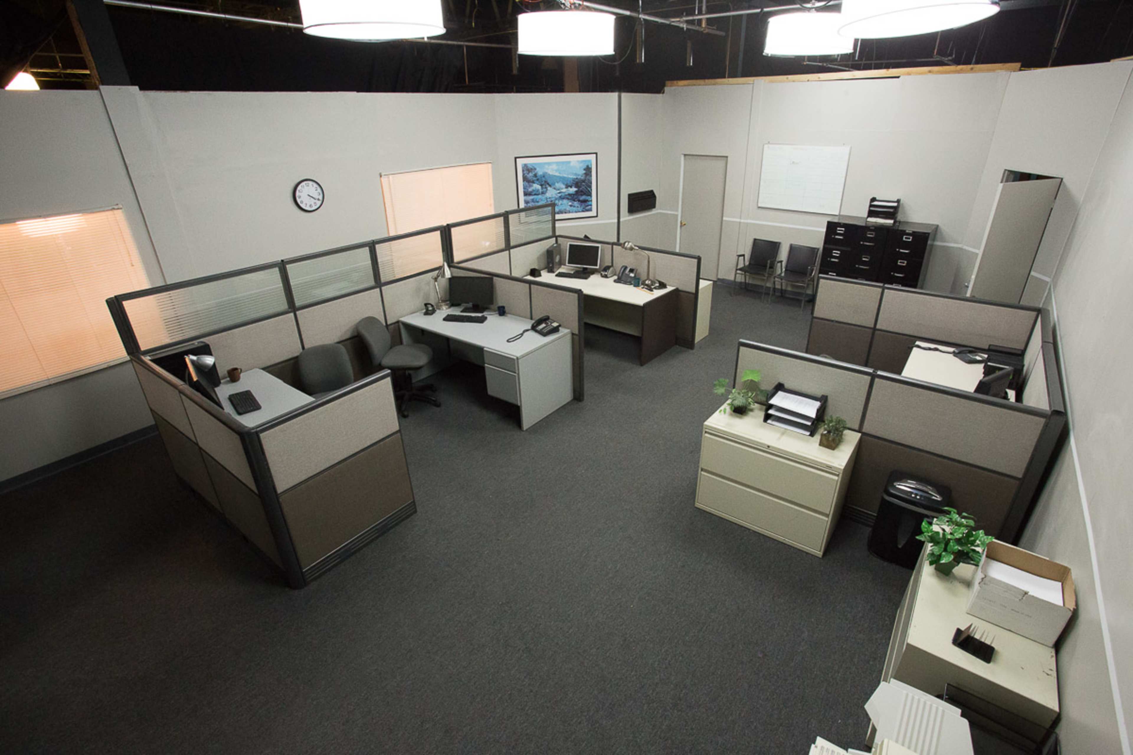 A modern office space featuring cubicles, desks with computers, and filing cabinets under overhead lights.