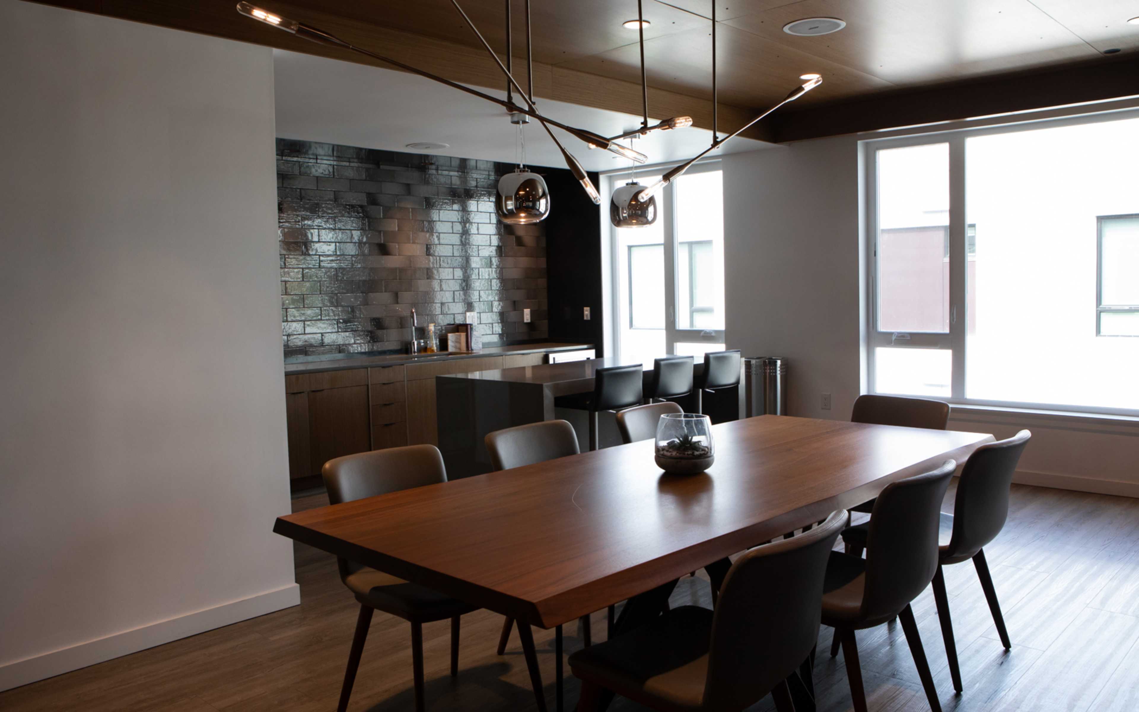 A modern dining area with a large wooden table surrounded by gray chairs, adjacent to a kitchen featuring dark tiles and cabinetry.