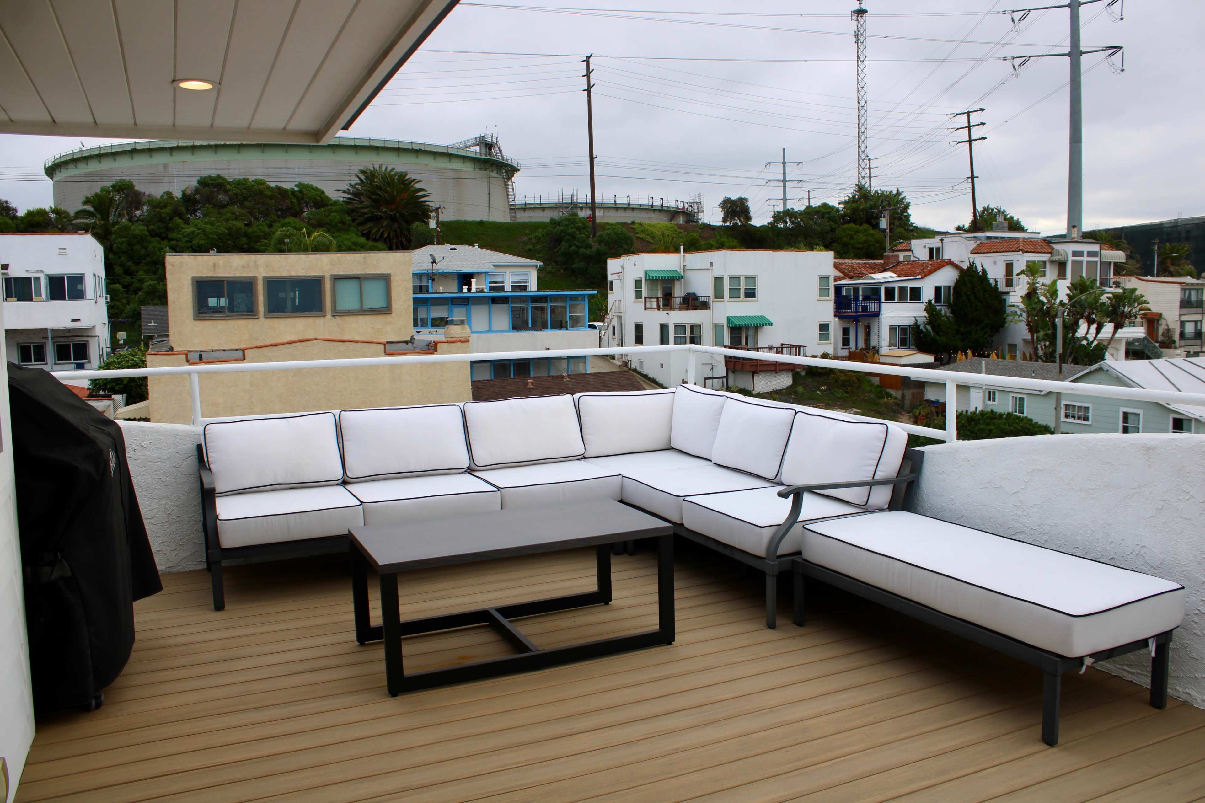 The image shows a modern outdoor seating area with a sectional couch and a coffee table on a deck, overlooking a neighborhood with various houses and a large tank in the background.
