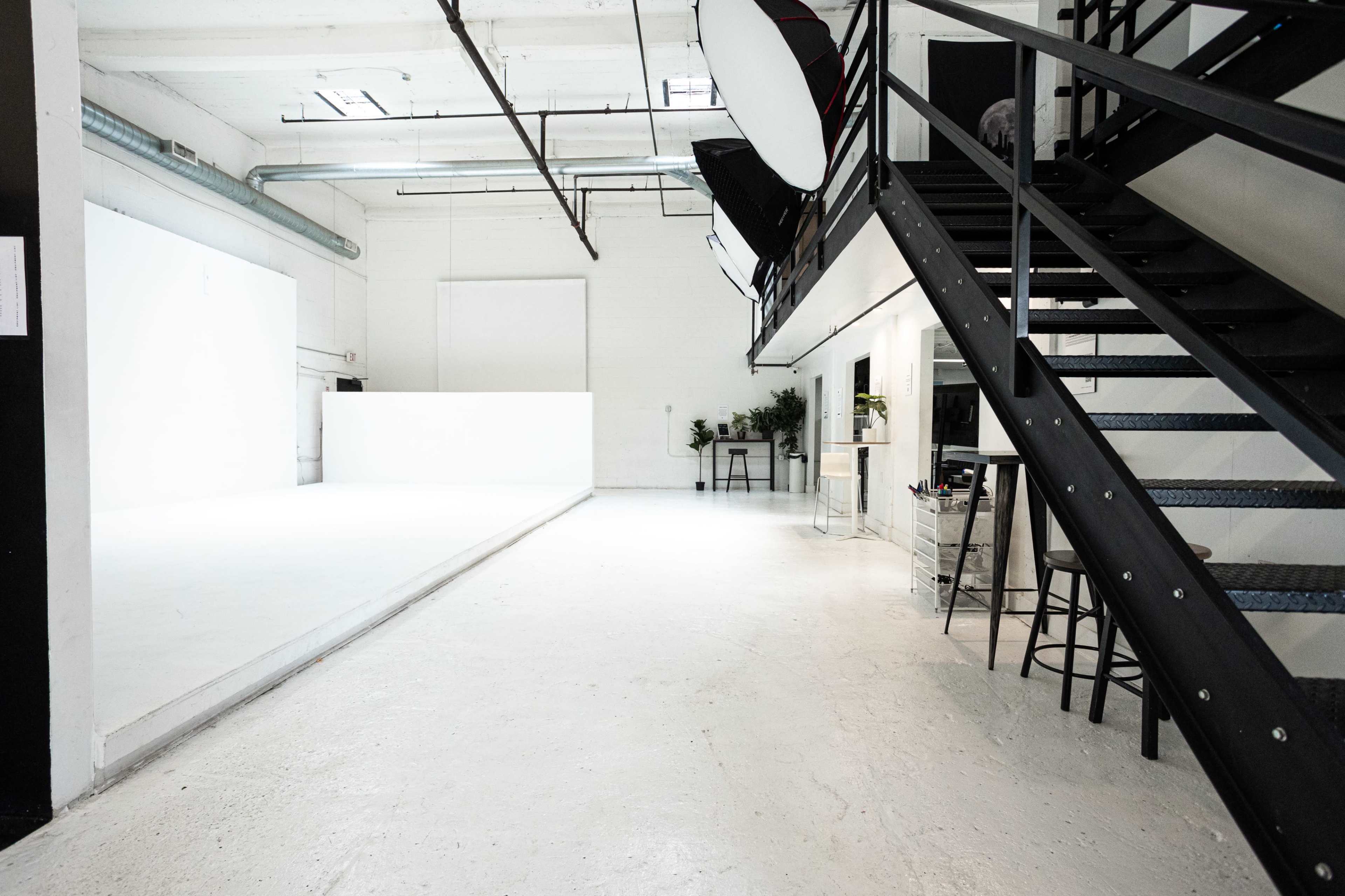 The image shows a spacious, well-lit studio with a large white backdrop, a staircase on the right, and some plants positioned near the back.