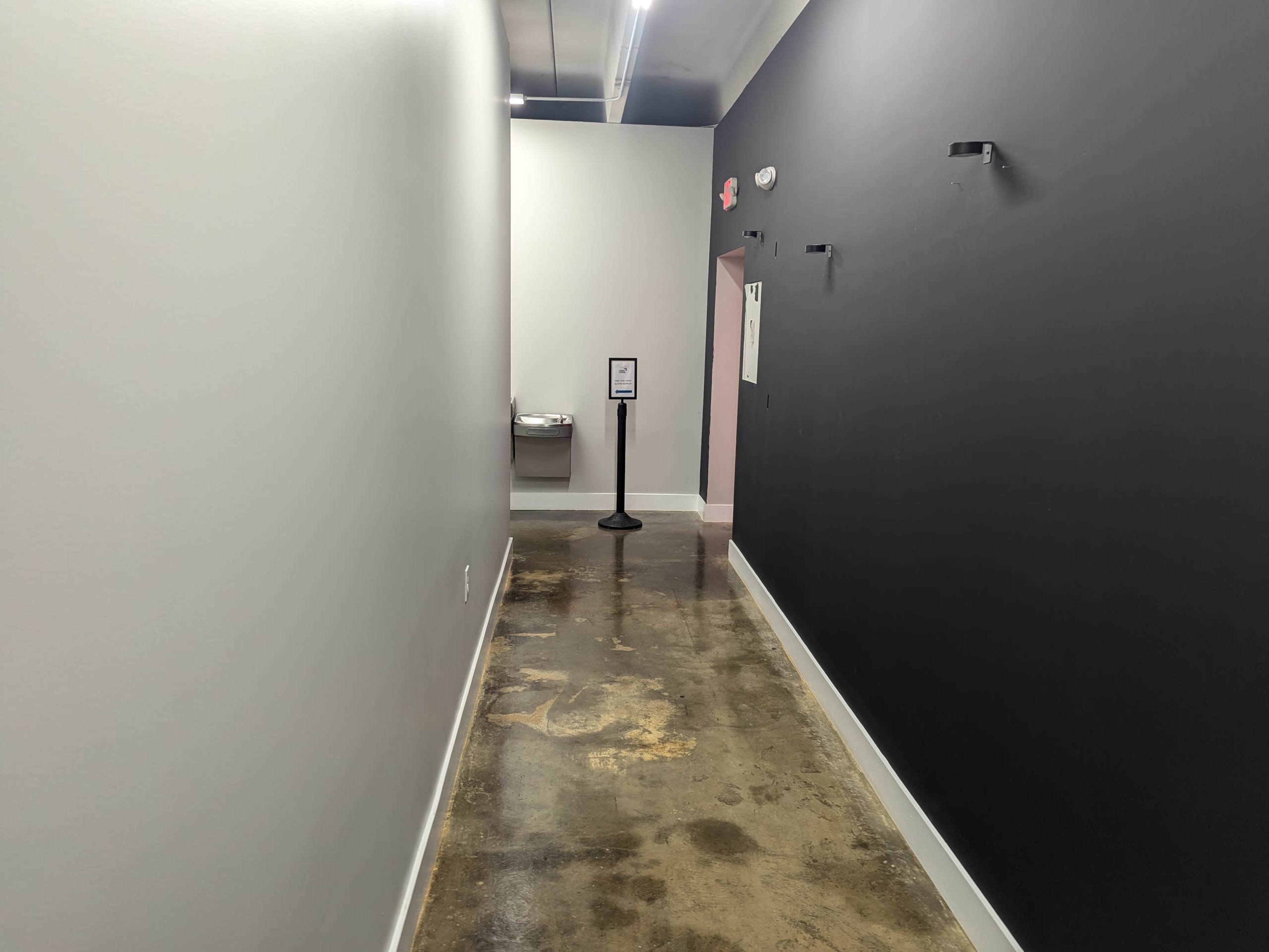 The image shows a narrow hallway with a gray wall on one side and a black wall on the other, leading to a pink doorway at the end.