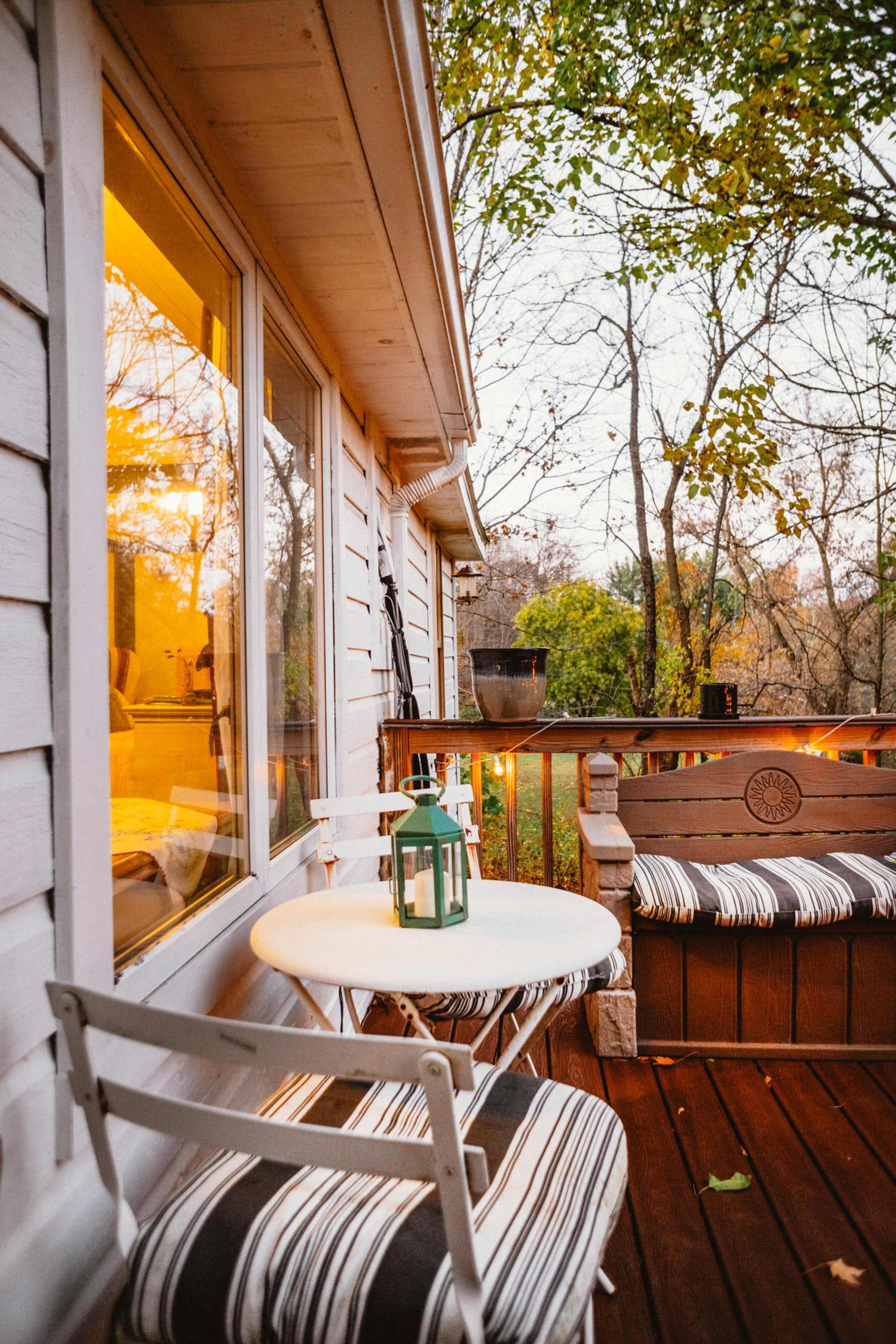 A small outdoor seating area features a round table with a green lantern and two striped chairs next to a wooden bench, surrounded by trees.