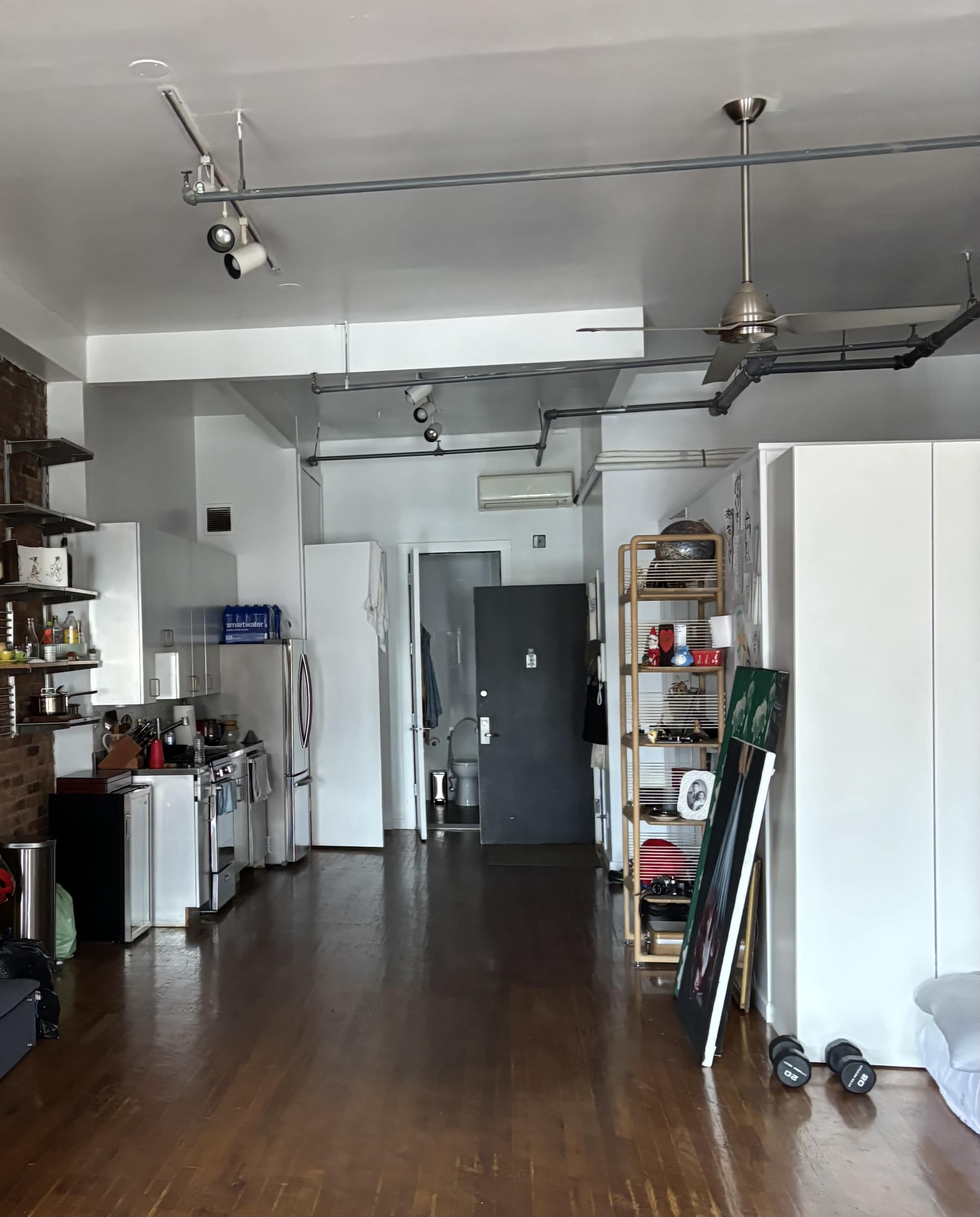 The image shows an open living space featuring a kitchen area with a stove and shelves, a doorway at the far end, and various items scattered around, including a large painting and exercise equipment.