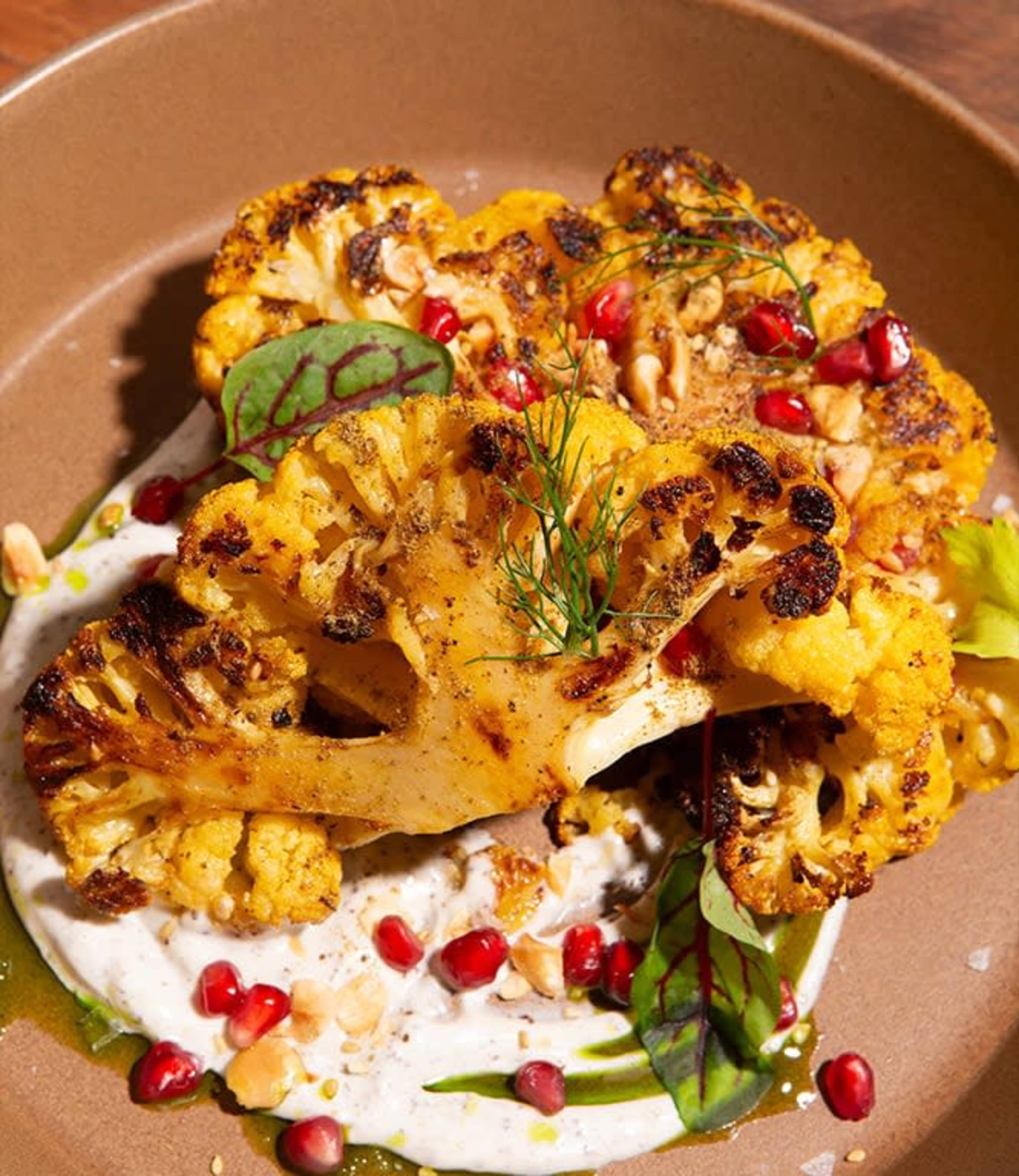 A platter features roasted cauliflower steaks garnished with pomegranate seeds, herbs, and drizzled with a yogurt sauce.