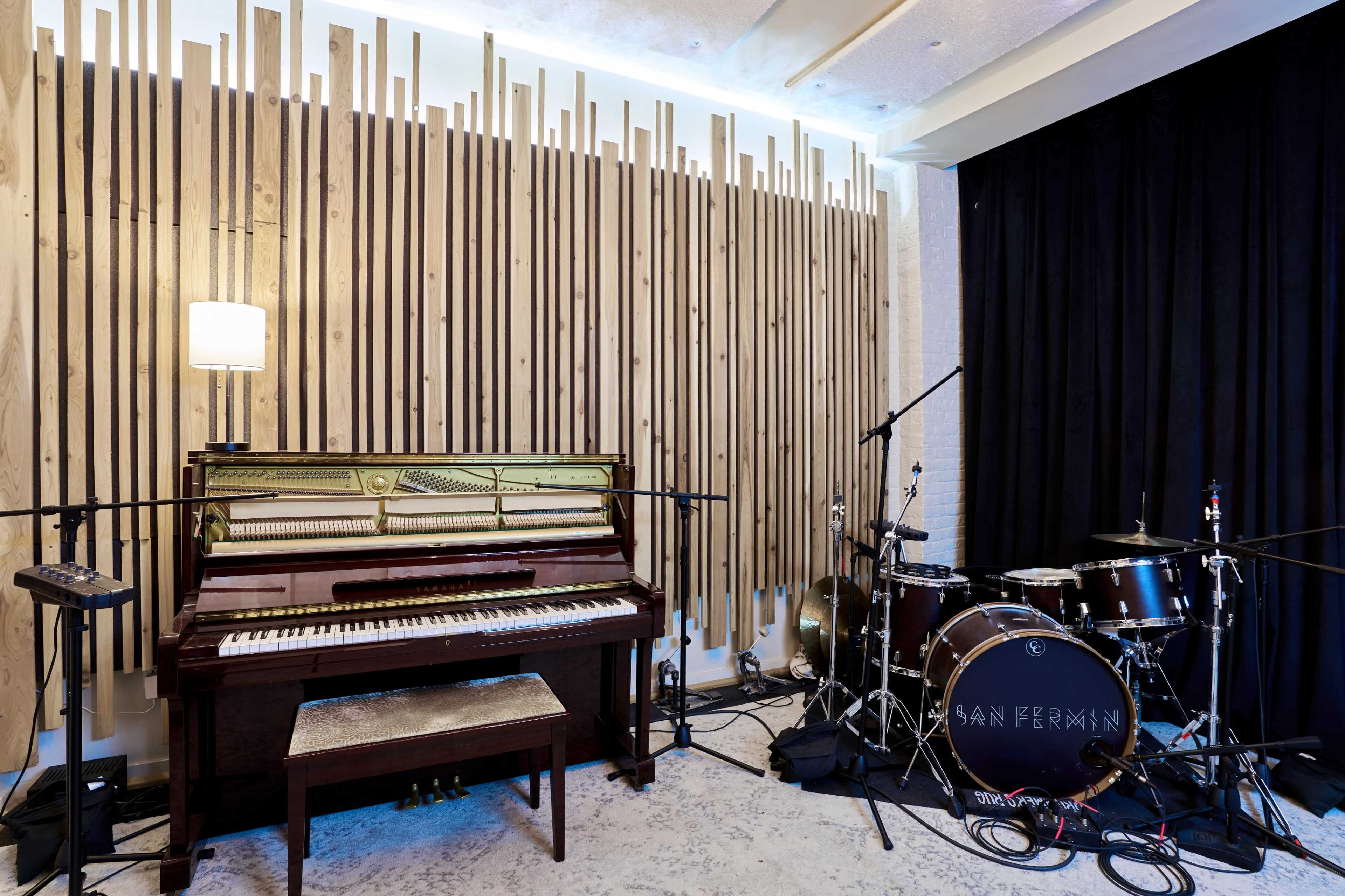 A vintage piano is positioned next to a drum set in a music studio with wooden accents on the walls and soft lighting.