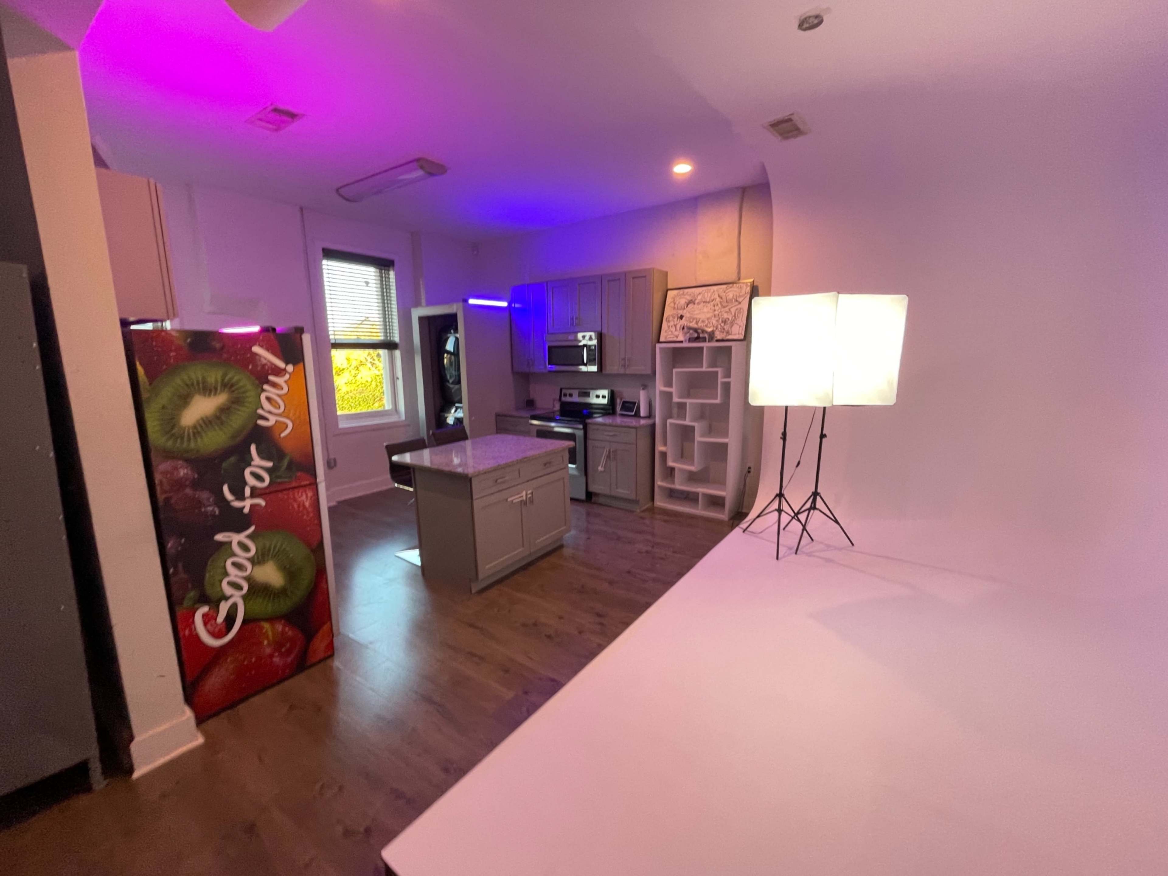 A modern kitchen with a central island, a colorful refrigerator on one side, and soft lighting fixtures set up for a photoshoot.