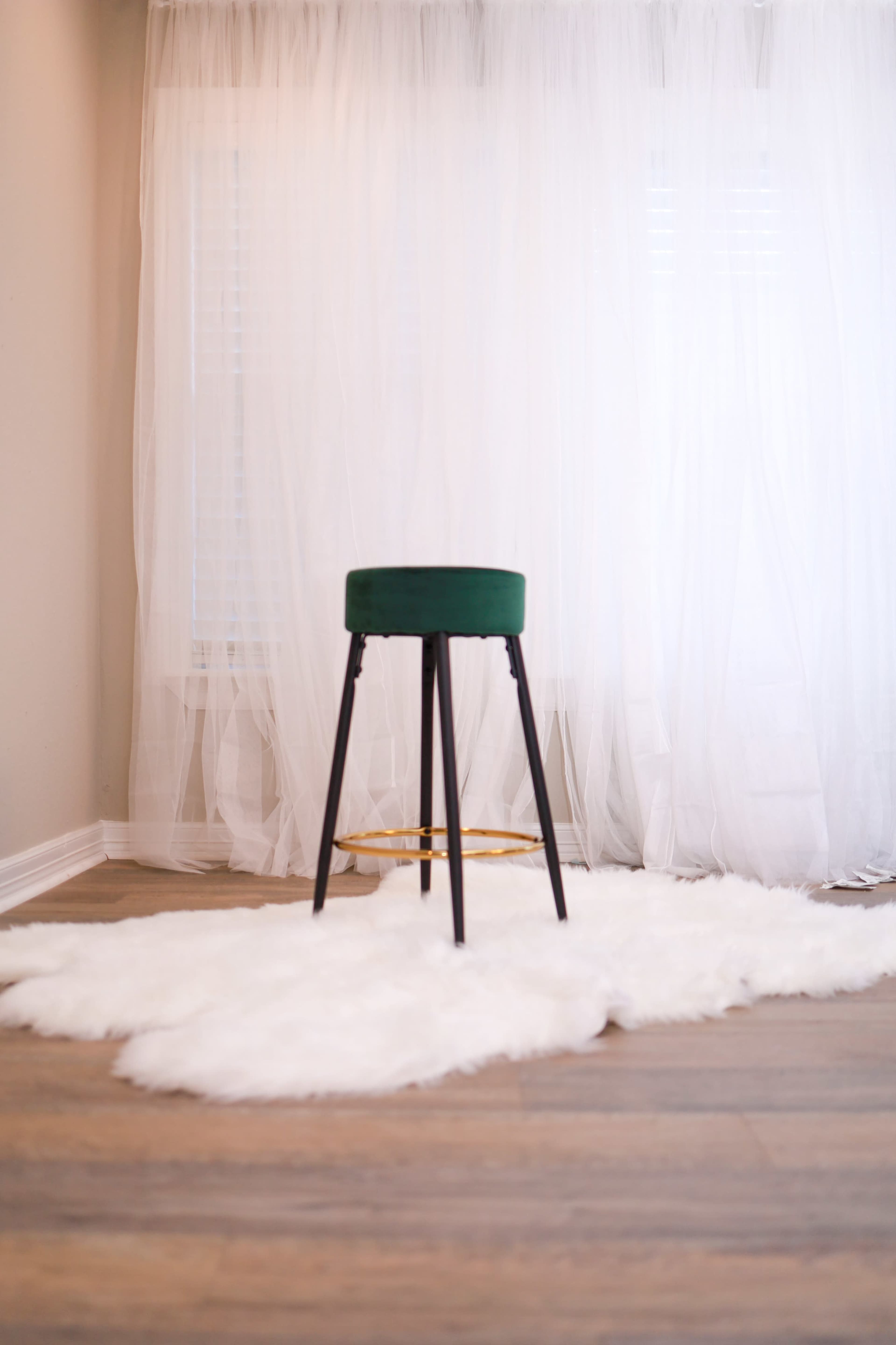 A green stool with black legs stands on a white faux fur rug in front of sheer white curtains.