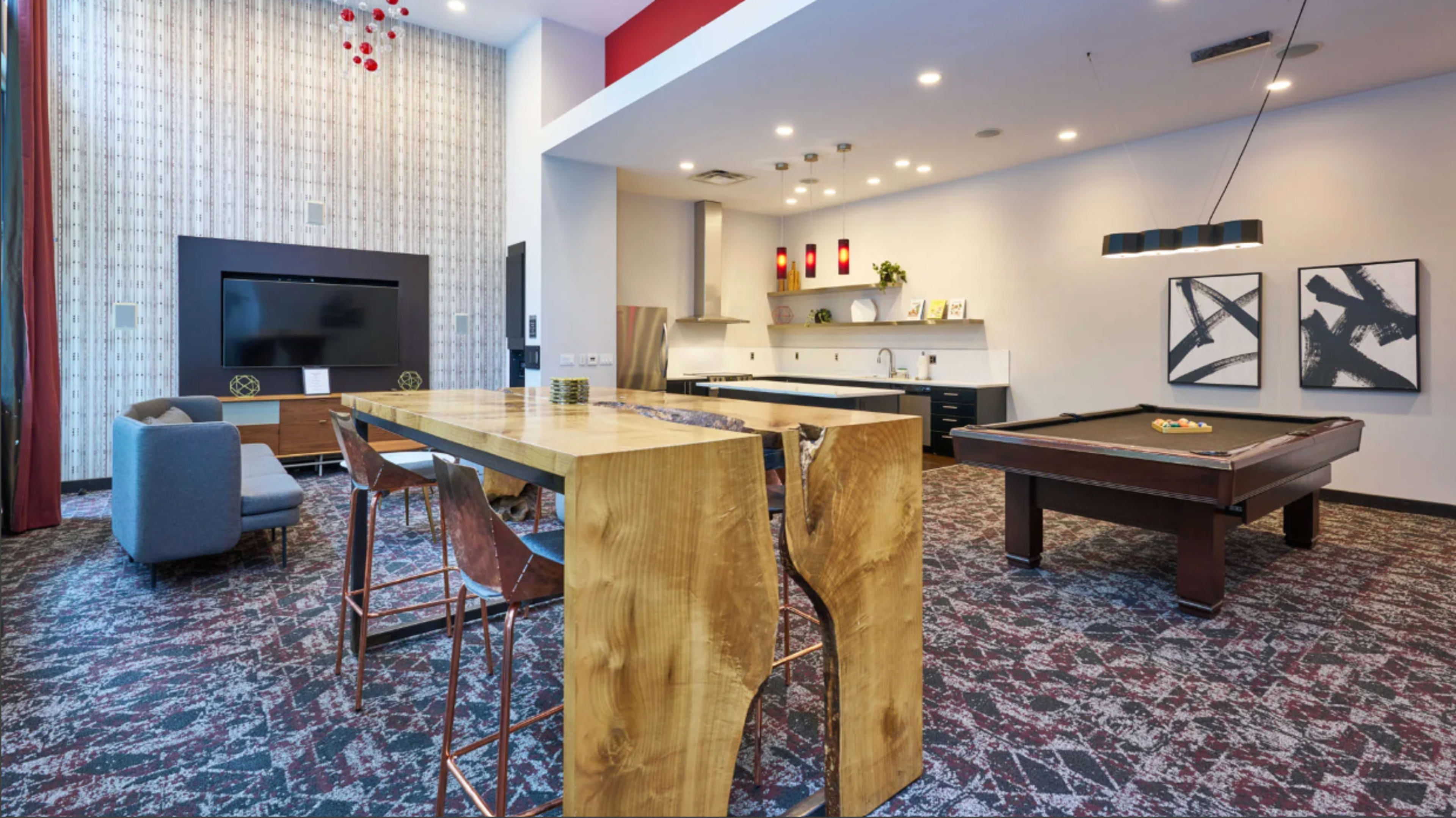 The image shows a modern communal lounge area featuring a large wooden table, a pool table, a kitchen space, and a seating arrangement with a television on the wall.