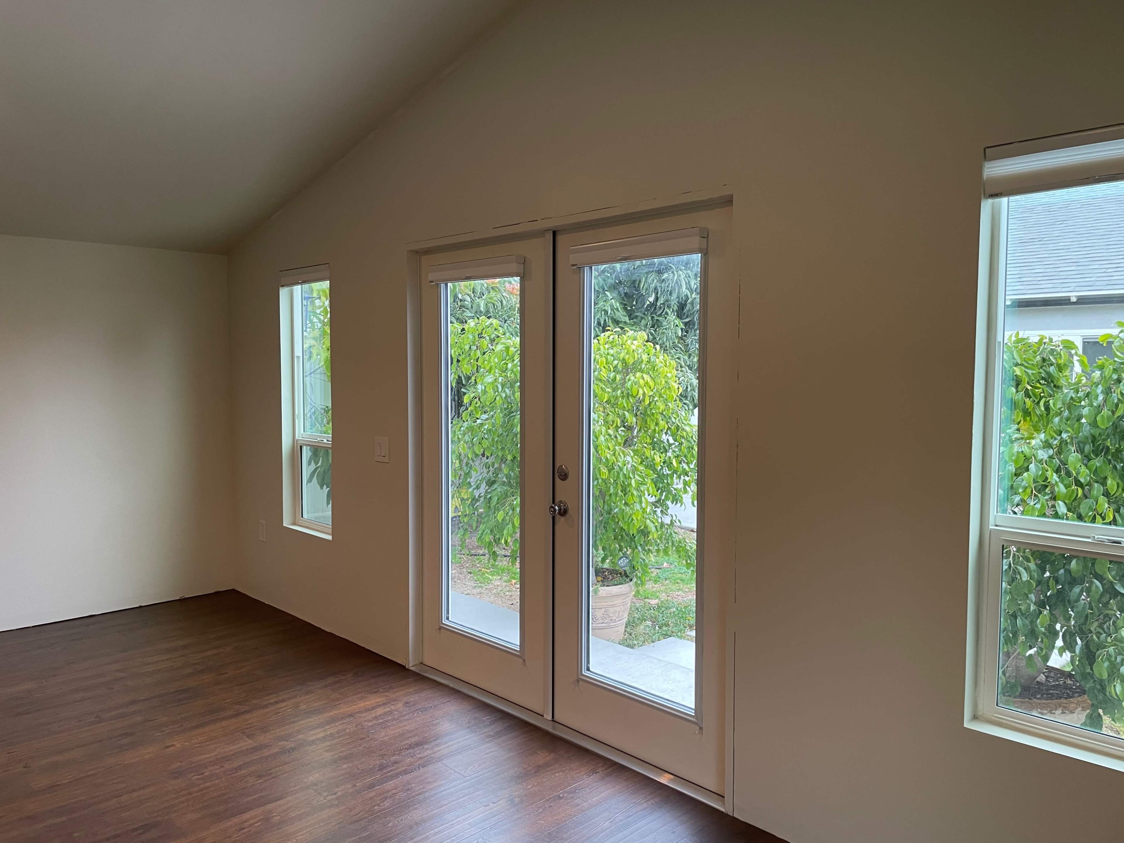 An interior room with two large windows and a set of double doors leading to an outdoor space filled with greenery.