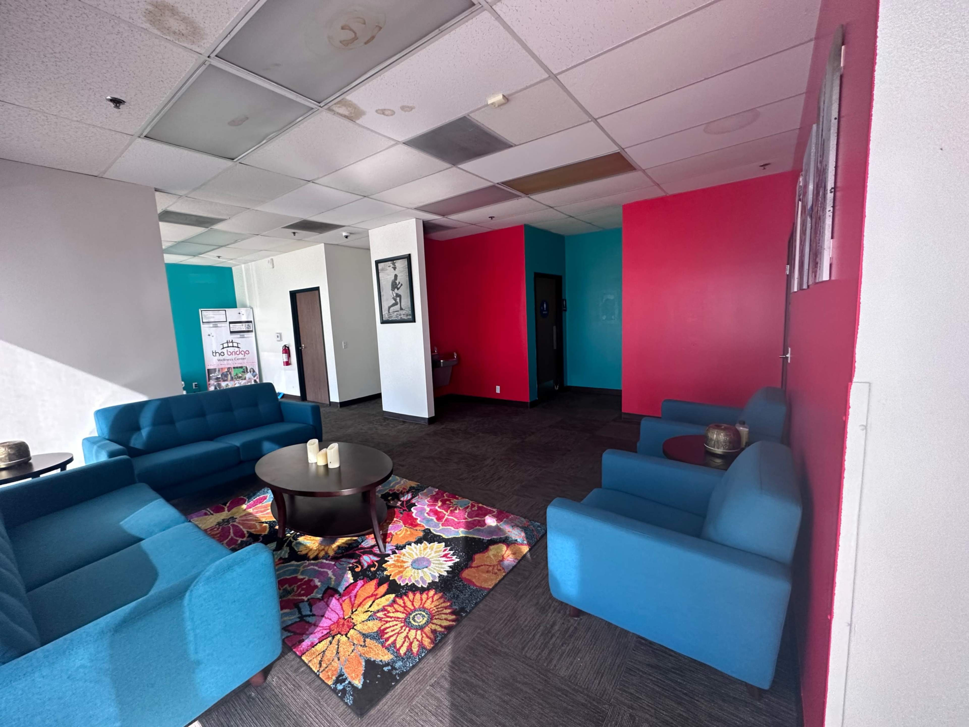 The image shows a waiting area with blue sofas, a round table, and a colorful floral rug against a backdrop of pink and white walls.