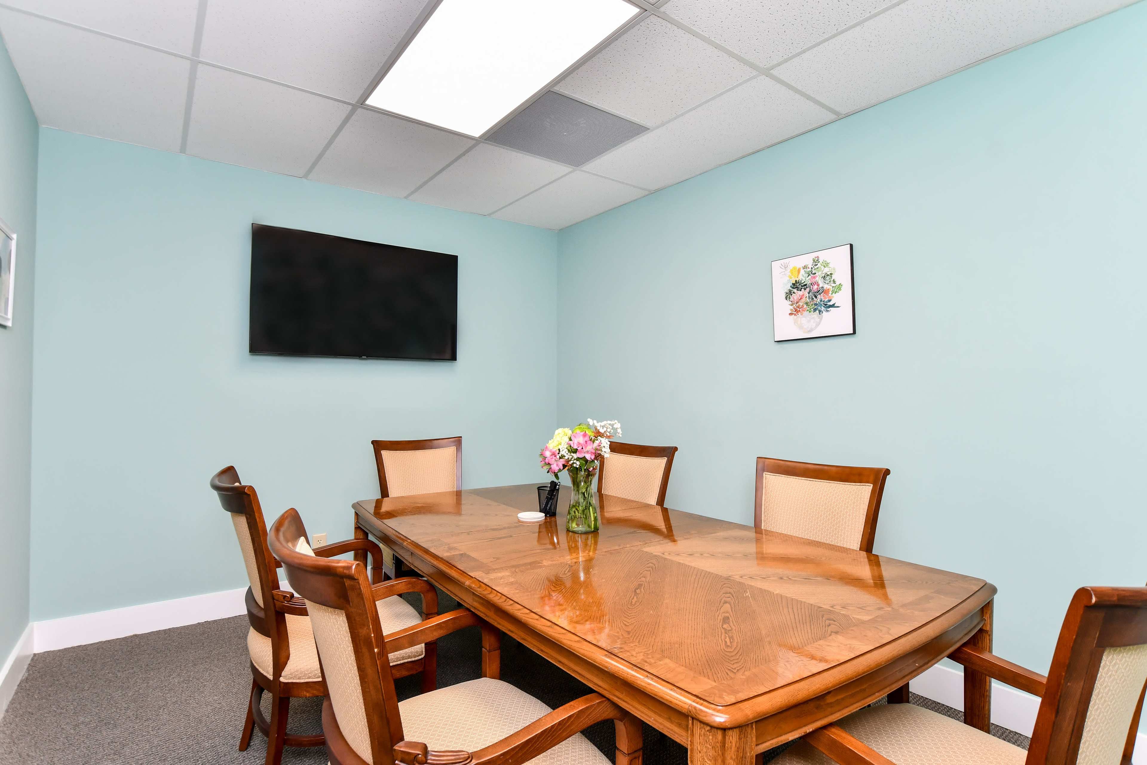 A wooden conference table is surrounded by six chairs in a light blue meeting room, featuring a wall-mounted television and a floral centerpiece.