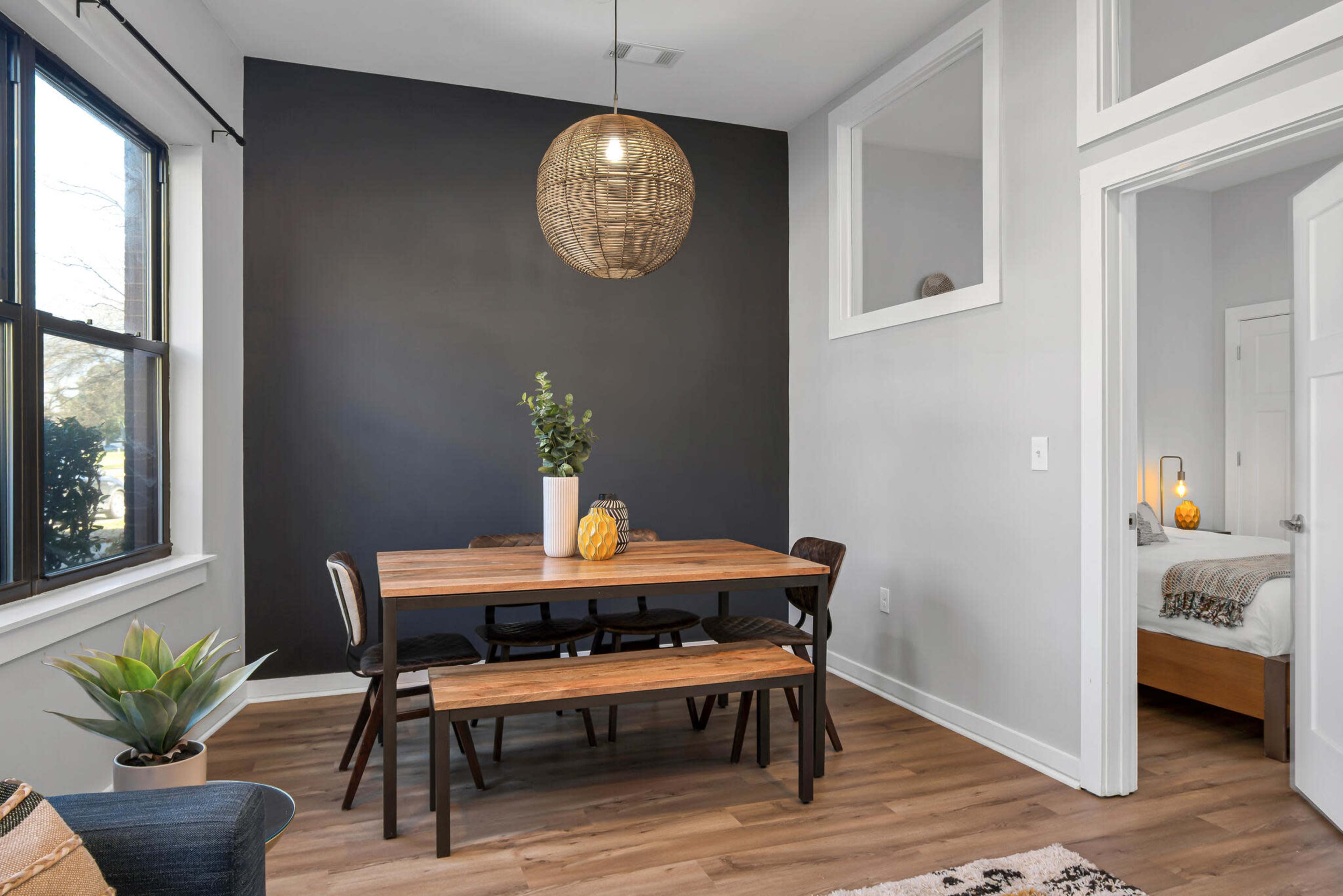 A dining area features a wooden table with black chairs, a pendant light, and a potted plant, adjacent to a bedroom with a view through an open doorway.
